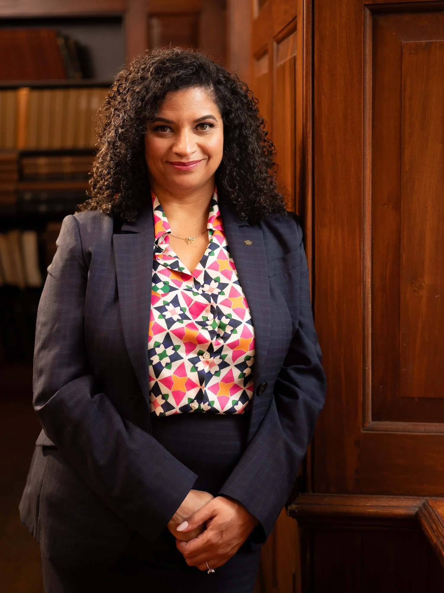 A woman with curly black hair wearing a dark blazer and a colorful geometric patterned shirt, standing in front of wooden cabinets in a room lined with books.