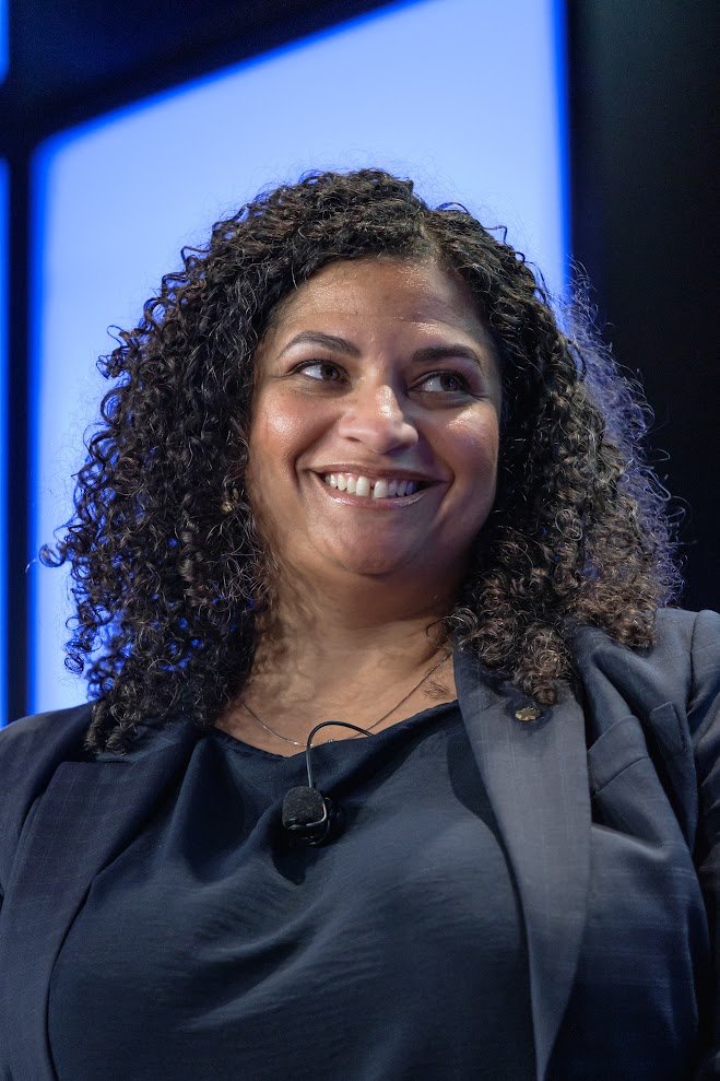 A woman with curly dark hair smiling, wearing a dark blazer and a microphone clipped to her shirt, in front of a blue modern background.