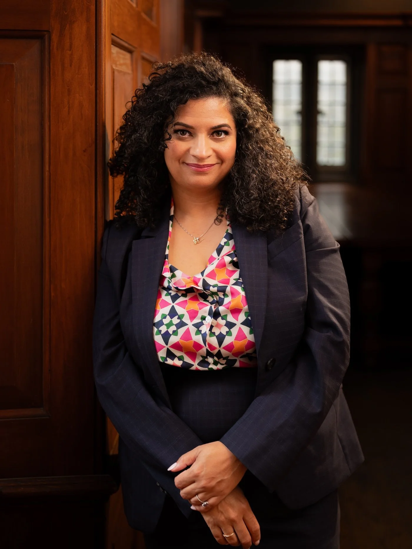 A woman with curly black hair, wearing a dark blazer and a colorful patterned blouse, standing in front of wooden paneling and windows.
