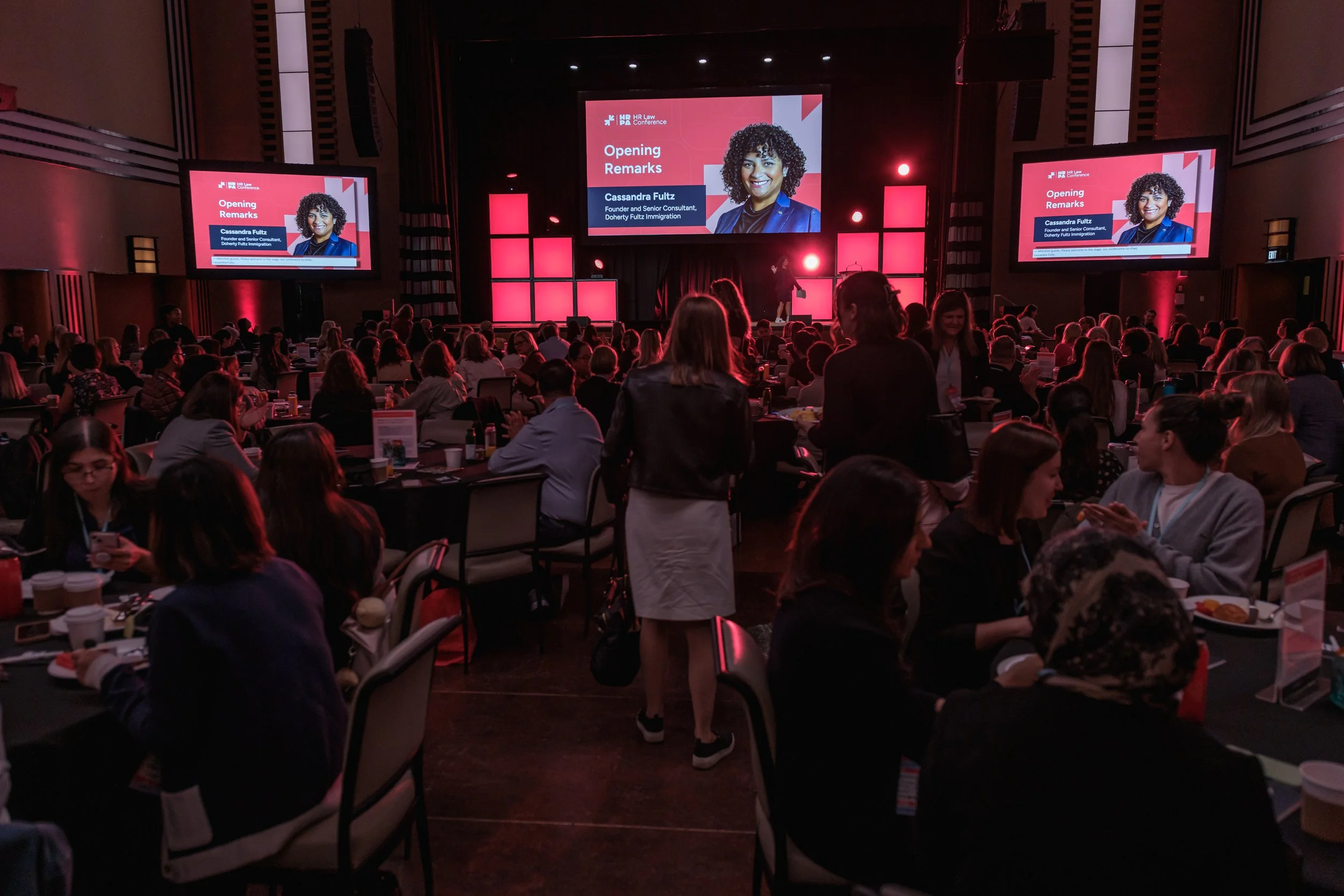 A large conference room filled with many seated attendees listening to a speaker at the front. There are three large screens displaying a presentation titled 'Opening Remarks' featuring a woman. The room has dim lighting with red accent lights.