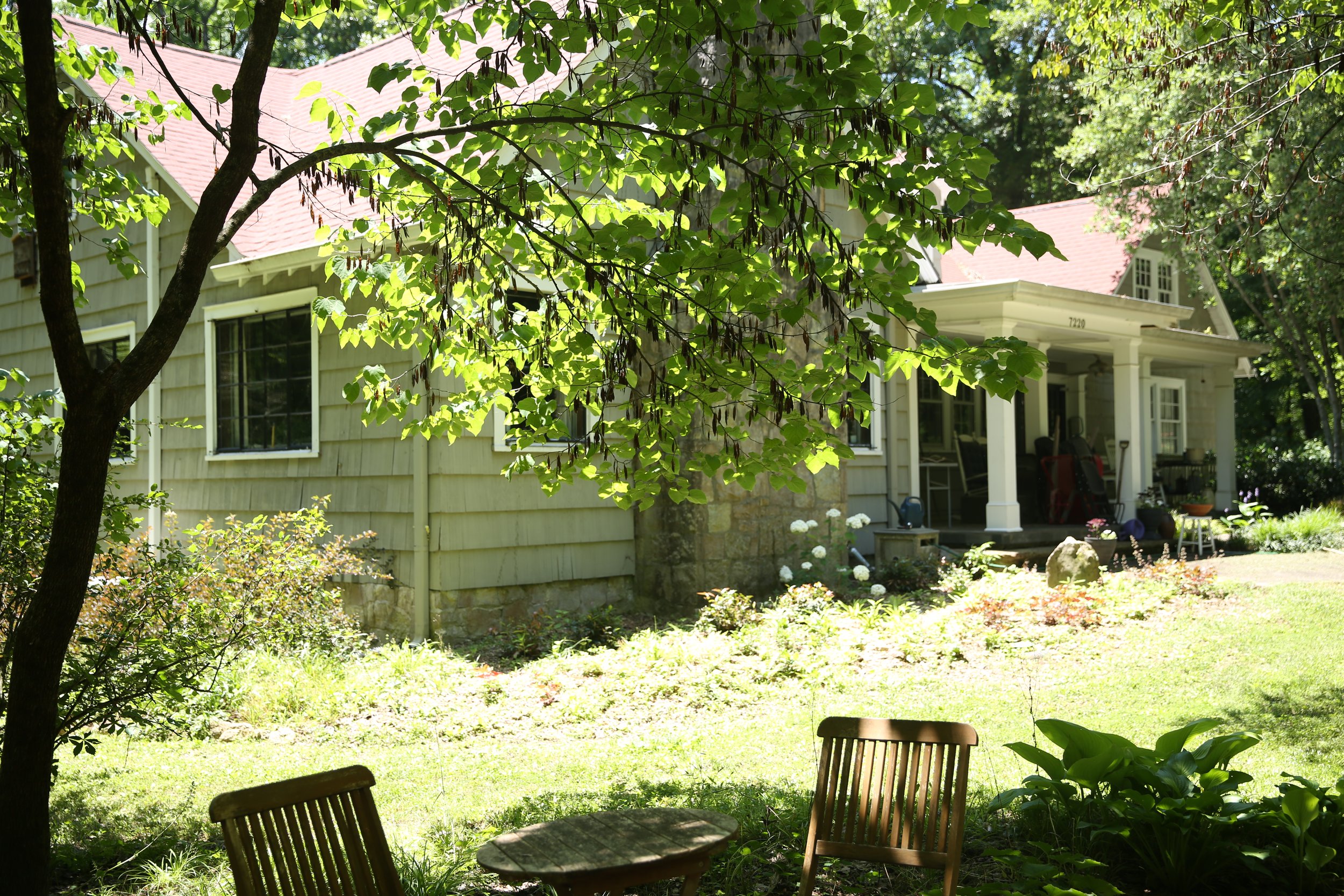 A backyard garden with wooden chairs and an overhanging tree with green leaves, neighboring a house with a porch and multiple windows, surrounded by lush greenery and trees.