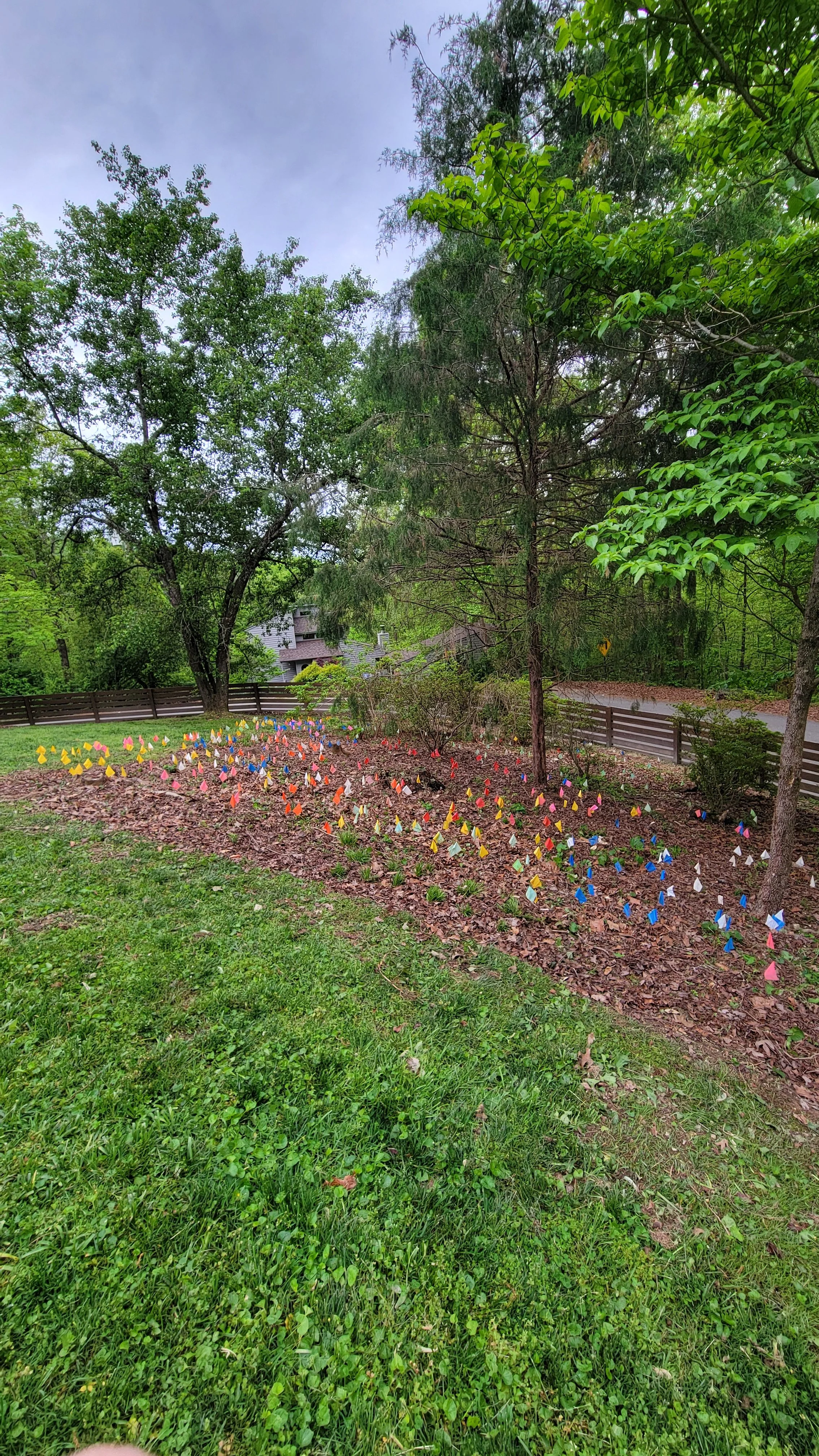 A garden bed with numerous small, colorful flags or paper triangles planted in the ground, surrounded by green grass and trees.