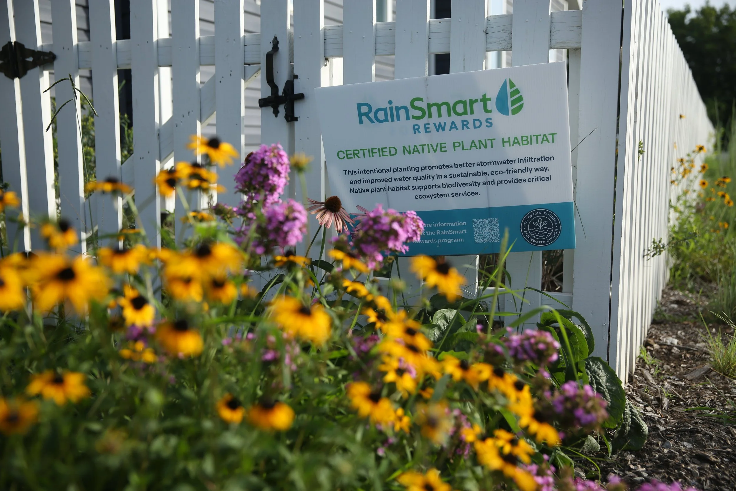 A white portable fence with a RainSmart Rewards sign promoting native plant habitat, surrounded by colorful flowers and greenery.