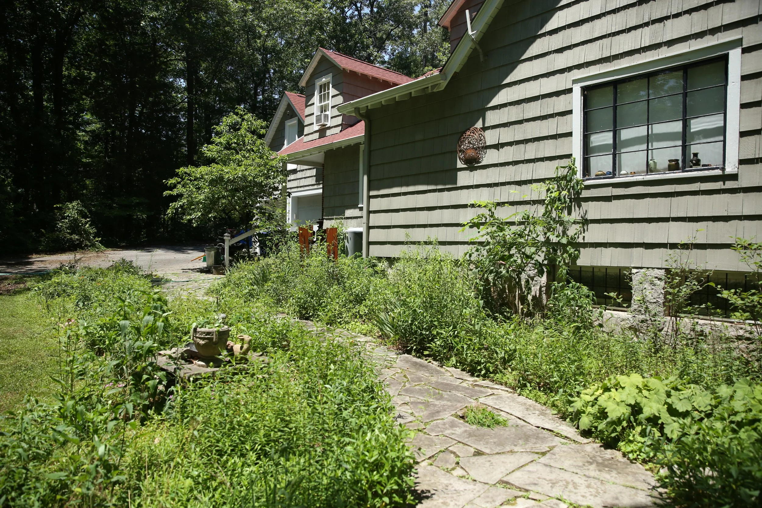 Side of a house with green siding, black framed window, decorated with pottery, set in a lush garden with a stone pathway and trees in the background.