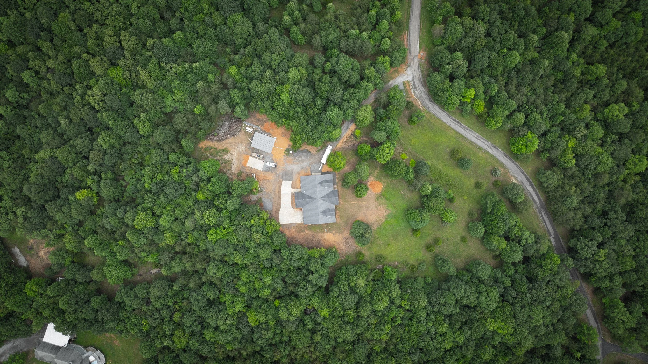 Aerial view of a house surrounded by dense green trees with a gravel driveway and some construction materials nearby.
