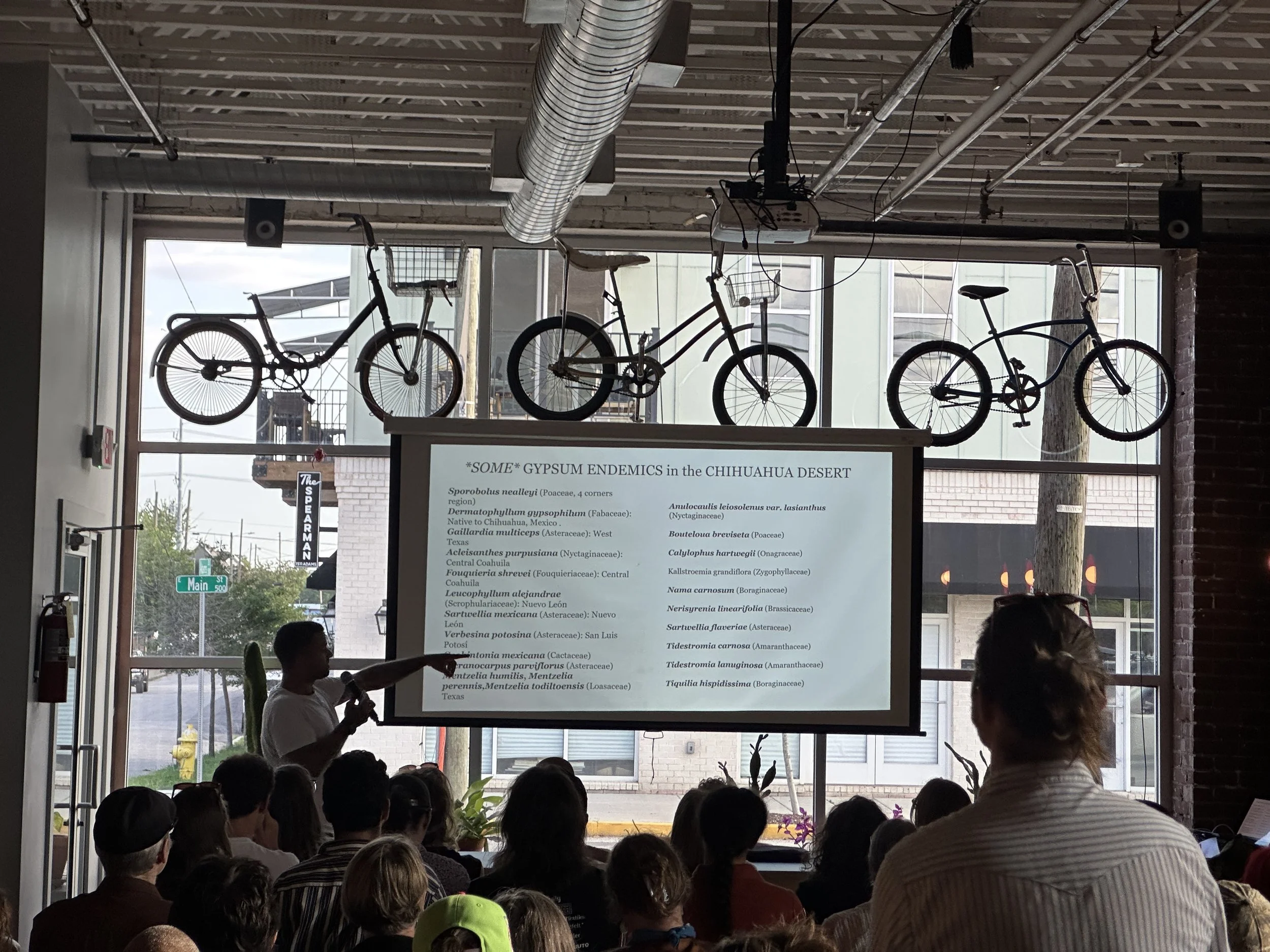 People attending a presentation in a room with large windows, displaying a projector screen with a list of plant species names.