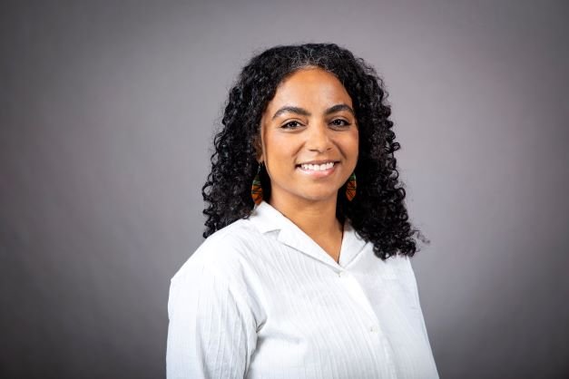 A woman with curly black hair smiling at the camera, wearing a white blouse and colorful earrings, against a gray background.
