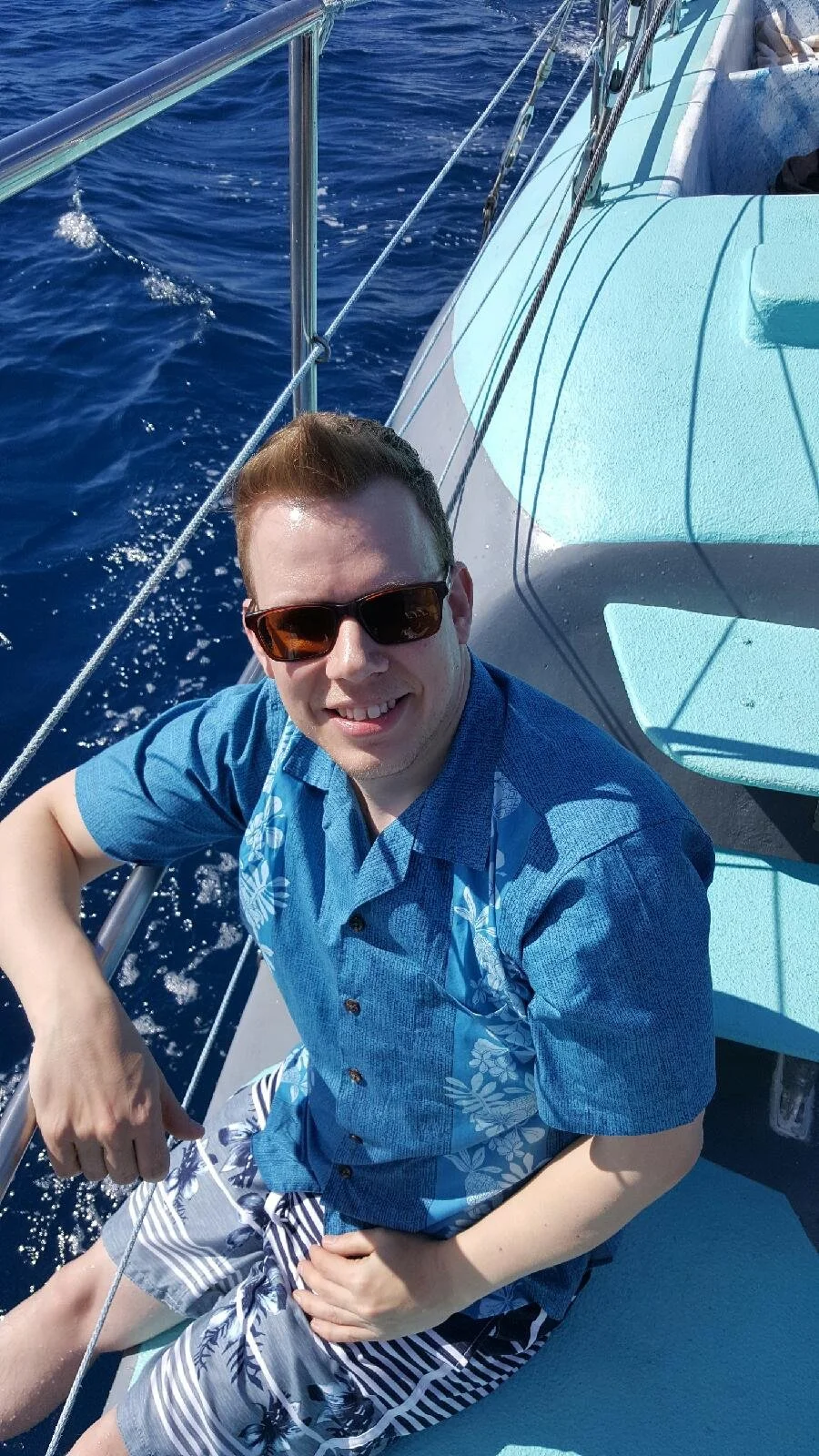 Man with sunglasses smiling on a sailboat, wearing a blue tropical shirt and striped shorts, over blue ocean water.