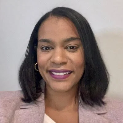 A woman with shoulder-length black hair, wearing a pink blazer and gold hoop earrings, smiling against a plain white background.