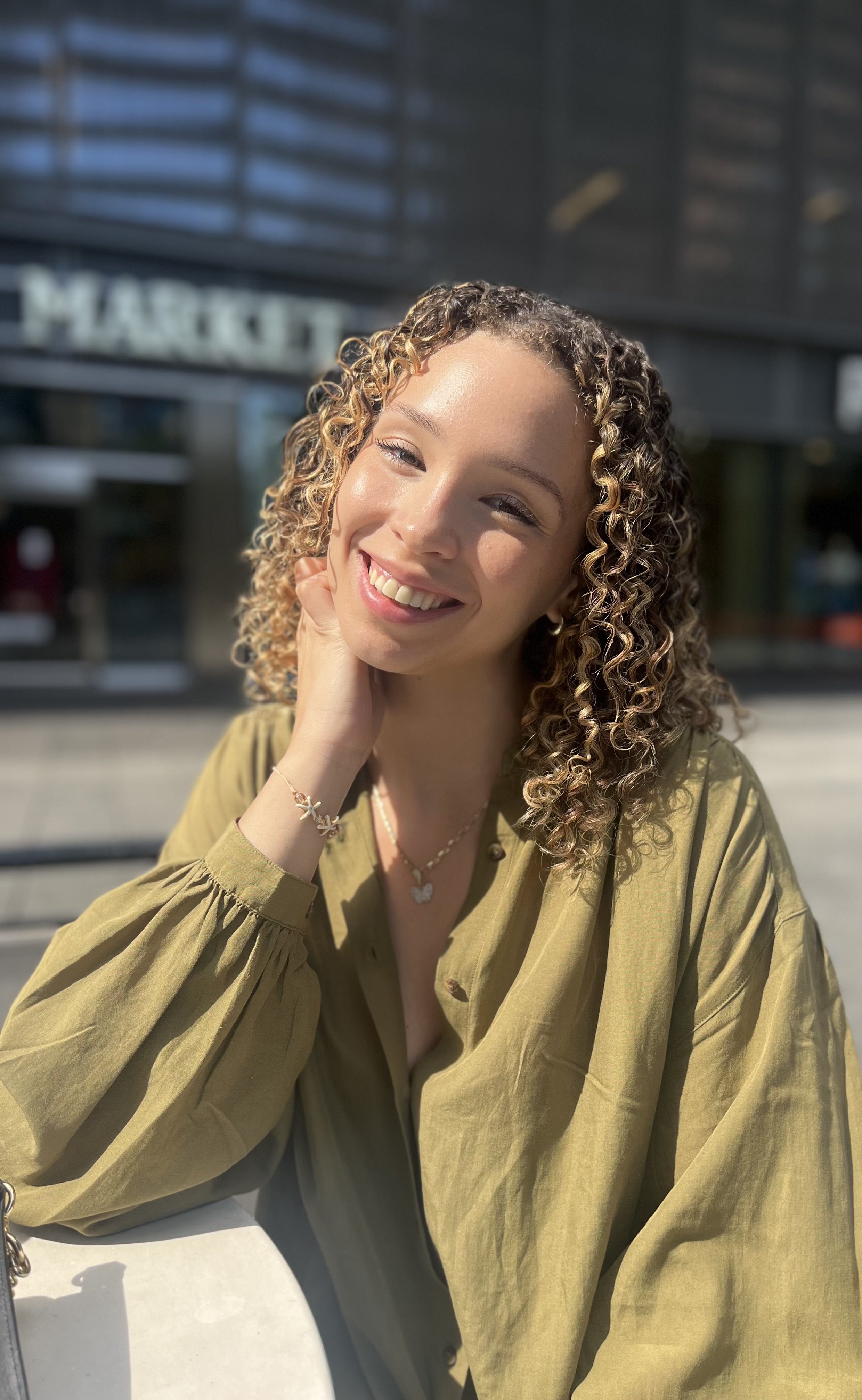 A young woman with curly blonde hair smiling outdoors, sitting at a table, wearing a green long-sleeved blouse and jewelry, with a modern building in the background.