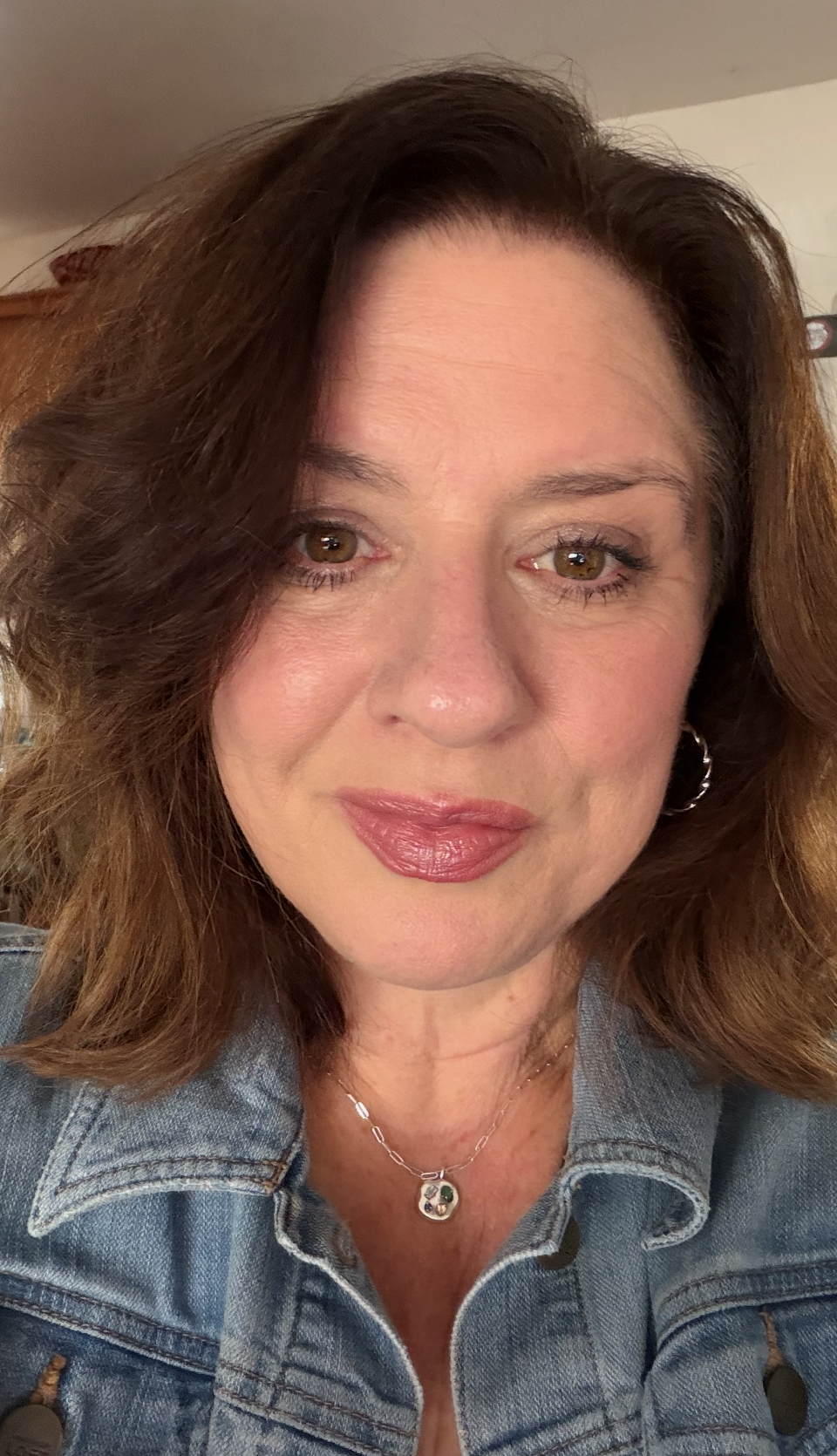 A woman with brown, wavy hair and light makeup, wearing a denim jacket and a silver necklace with a small pendant, smiling softly in a close-up portrait.
