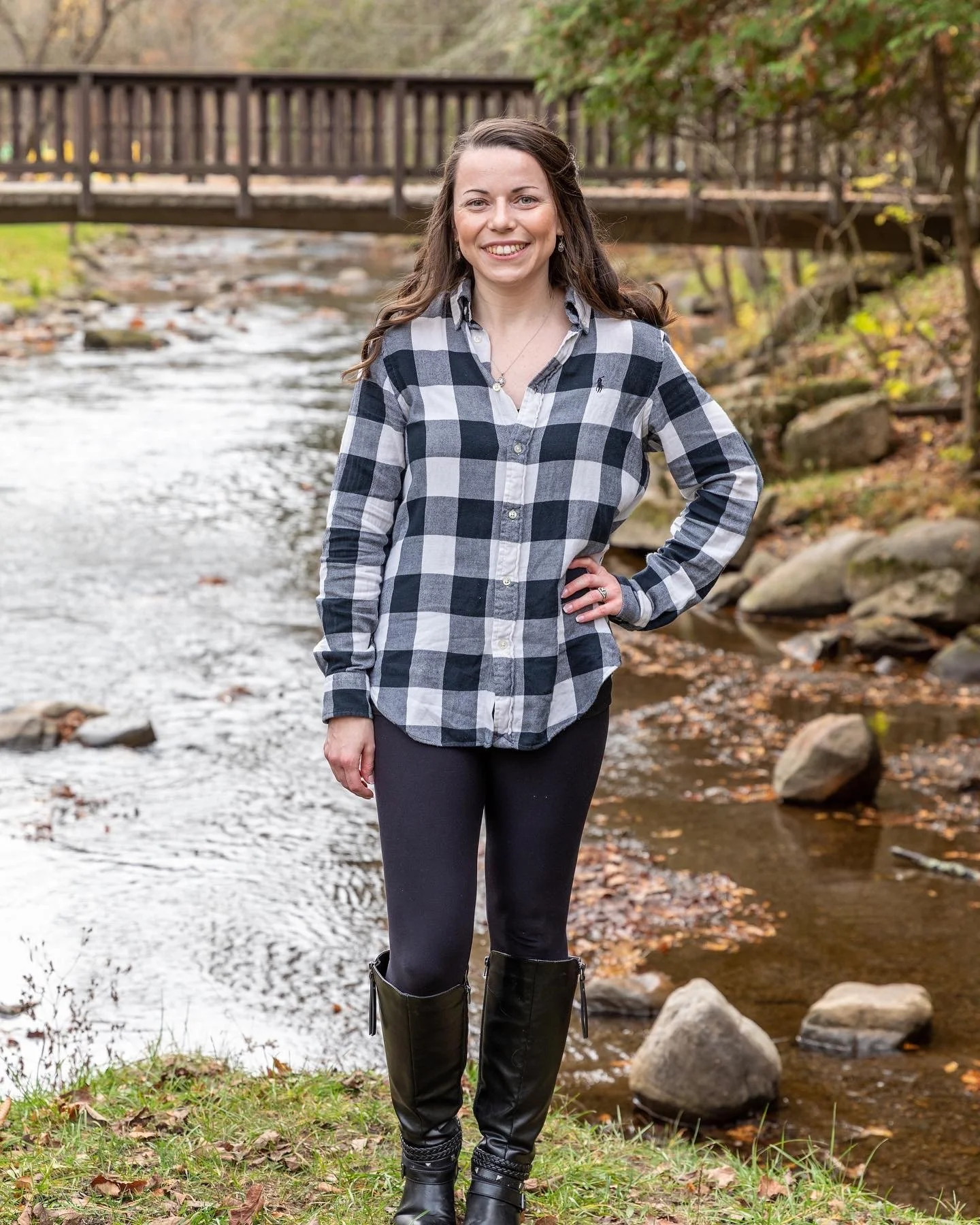 A woman standing by a creek with a wooden footbridge in the background, wearing a black and white checkered shirt, black leggings, and tall black boots, smiling at the camera.