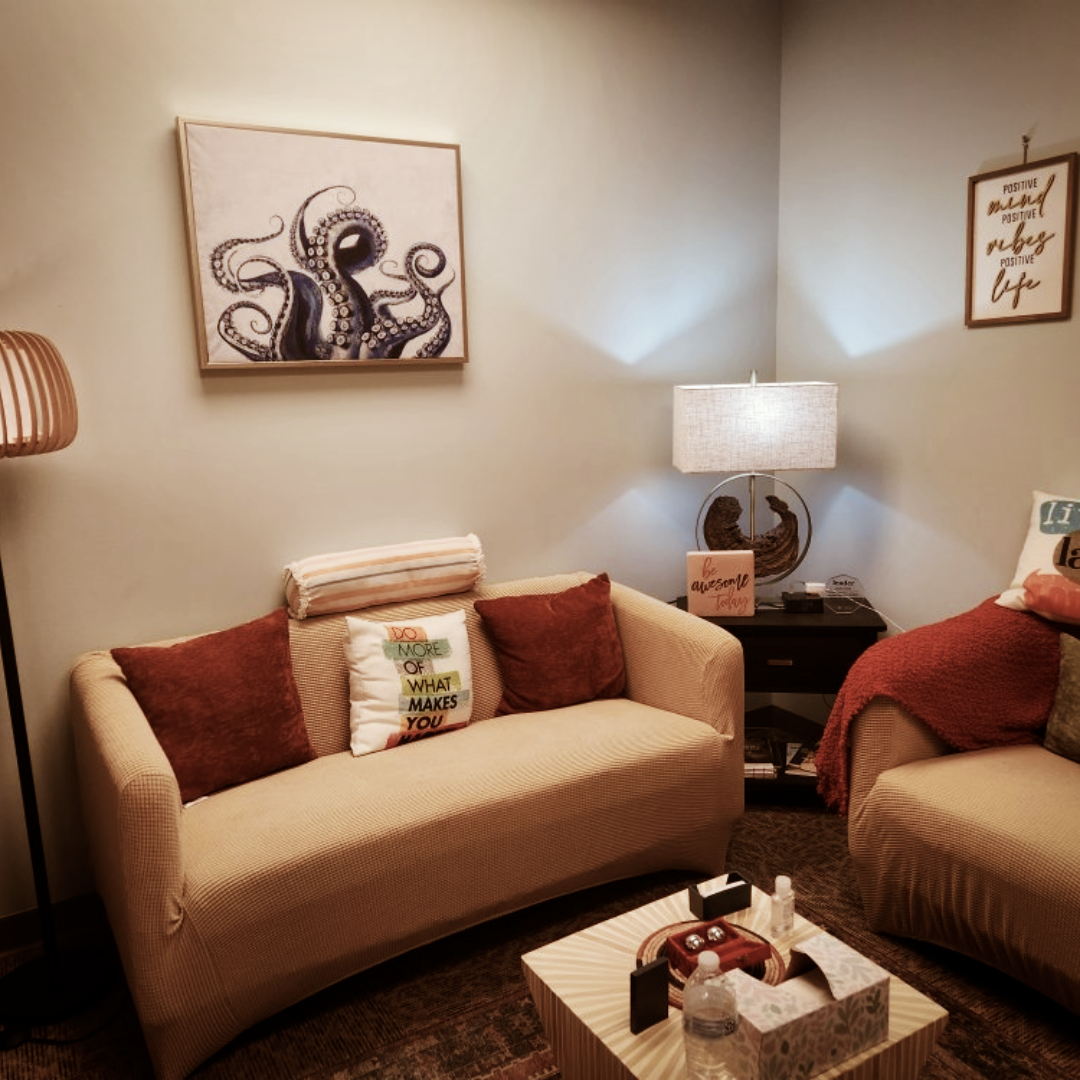 A cozy living room with two beige sofas, decorated with pillows, one with an inspirational quote. A wooden coffee table with items on it sits in front. A side table with a lamp and framed art, including an octopus painting, is in the corner. Wall decor includes motivational and positive message signs.