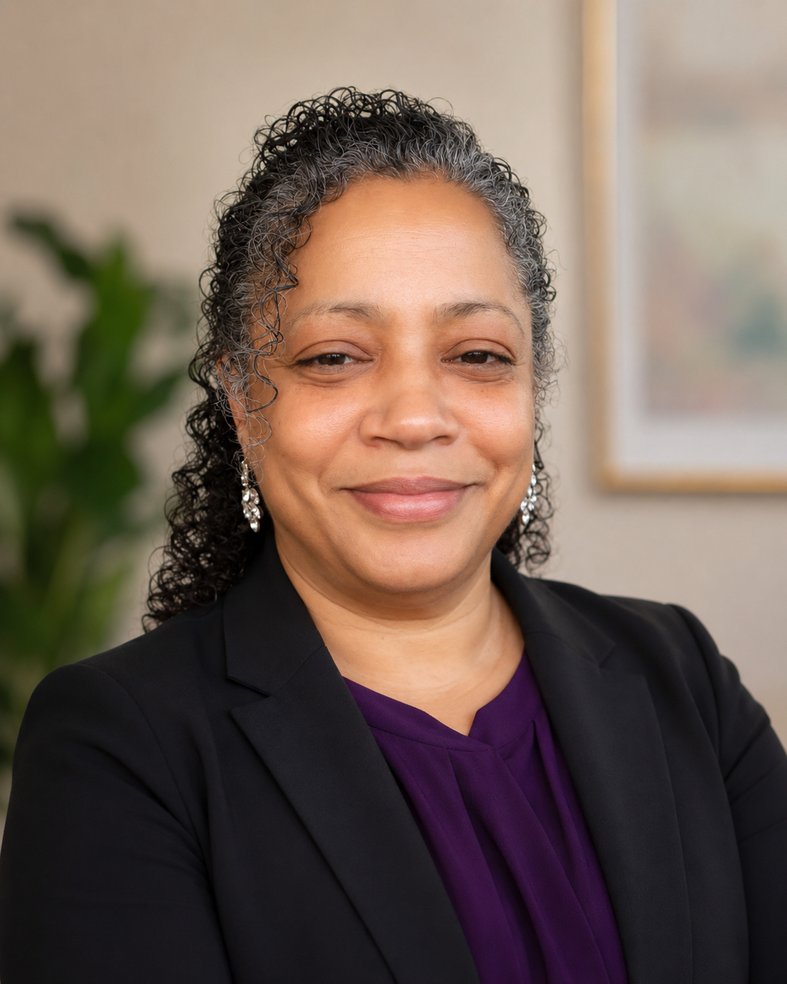 A professional woman with curly black and gray hair, wearing earrings, a purple blouse, and a black blazer, smiling in an office setting.