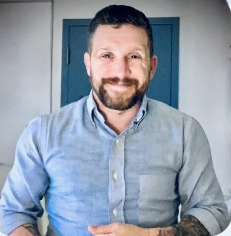 A man with a beard and short hair, wearing a light blue button-up shirt, smiling and sitting indoors in front of a blue door.