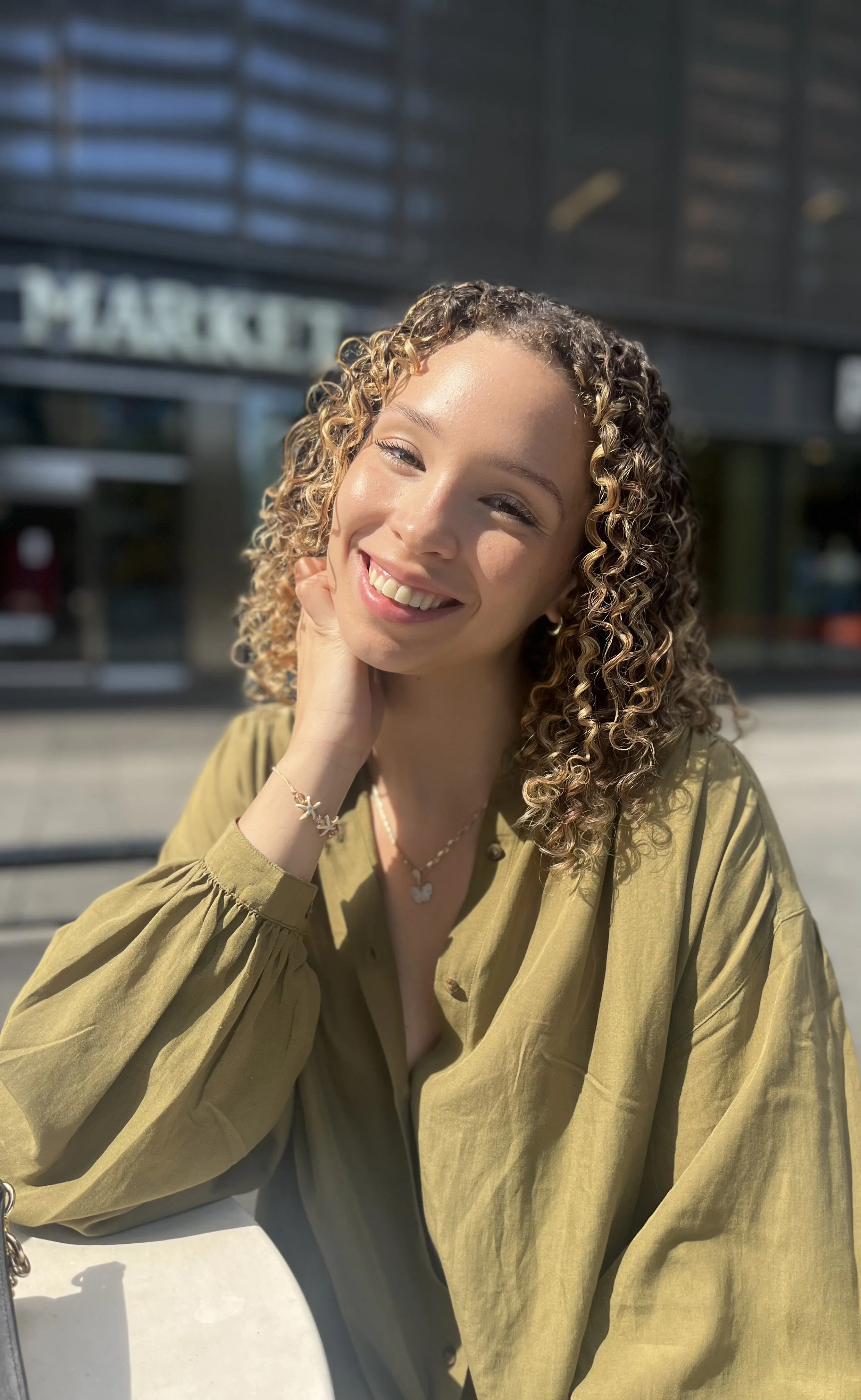 A young woman with curly blonde hair smiling and resting her face on her hand outdoors in sunlight, wearing a green top and jewelry, with a modern building in the background.