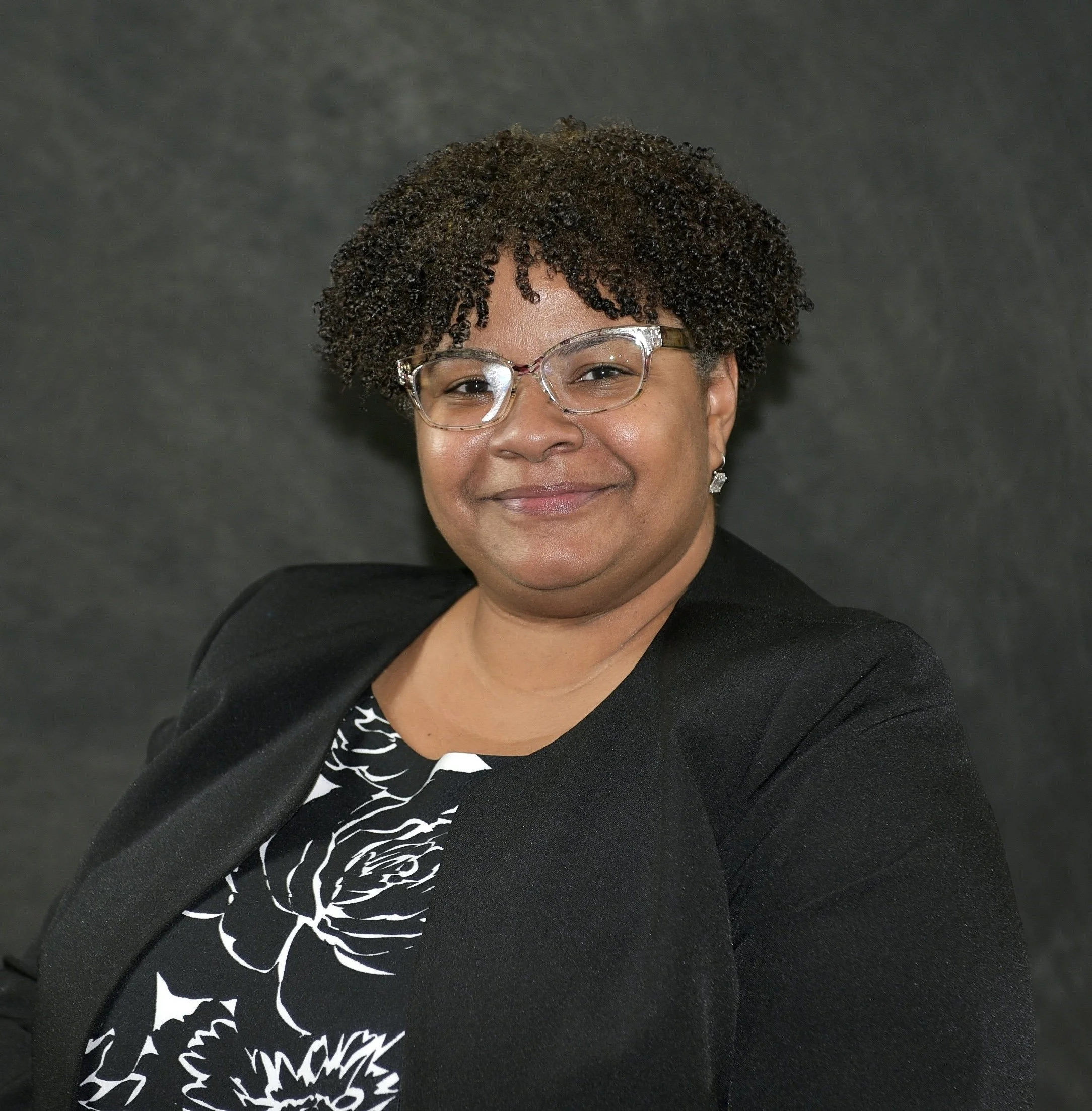 Portrait of a woman smiling, wearing glasses and a black blazer with a patterned top, against a dark, plain background.