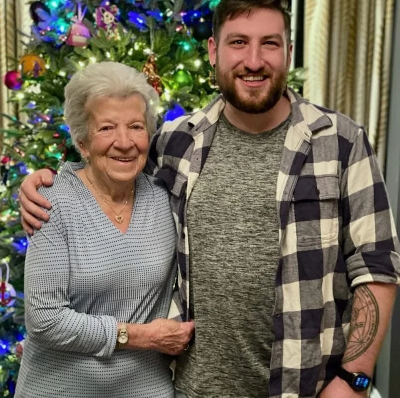 An elderly woman and a young man smiling and standing close together in front of a decorated Christmas tree with colorful lights and ornaments.