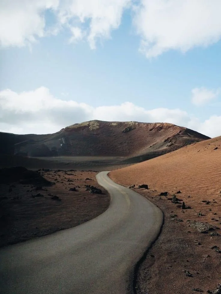 Camino serpenteante en un desierto con montañas al fondo y cielo con nubes.