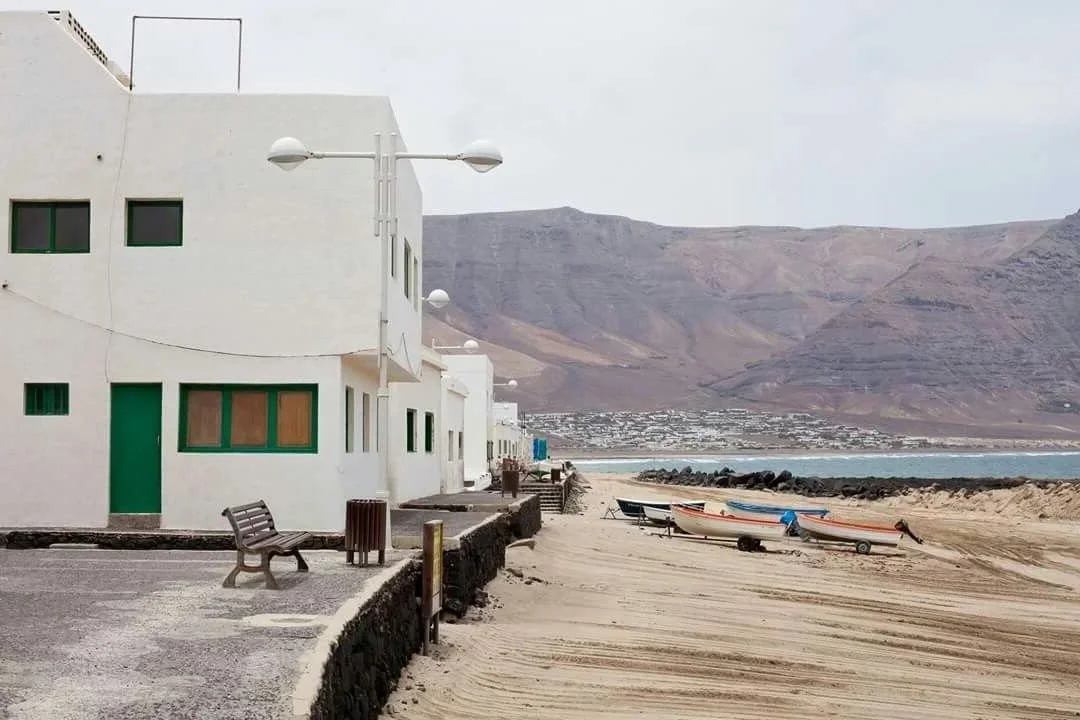 Casas blancas frente a la playa con barcos en la arena y montañas de fondo.