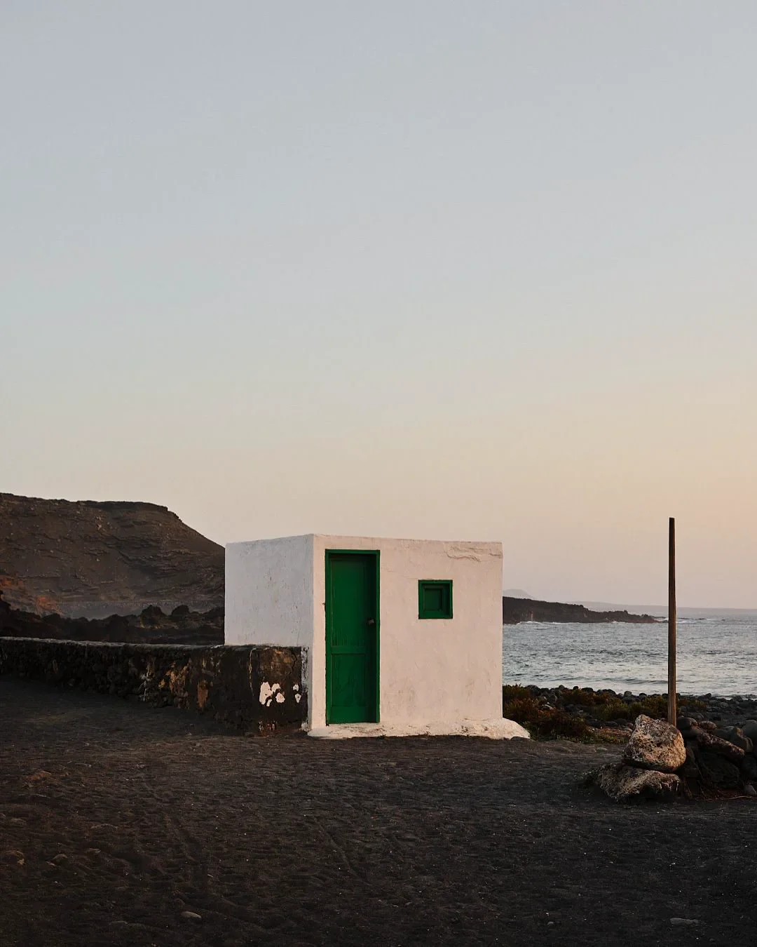 Una pequeña casa blanca con puerta y ventanas verdes junto a la playa, en un entorno natural, durante el atardecer o amanecer.