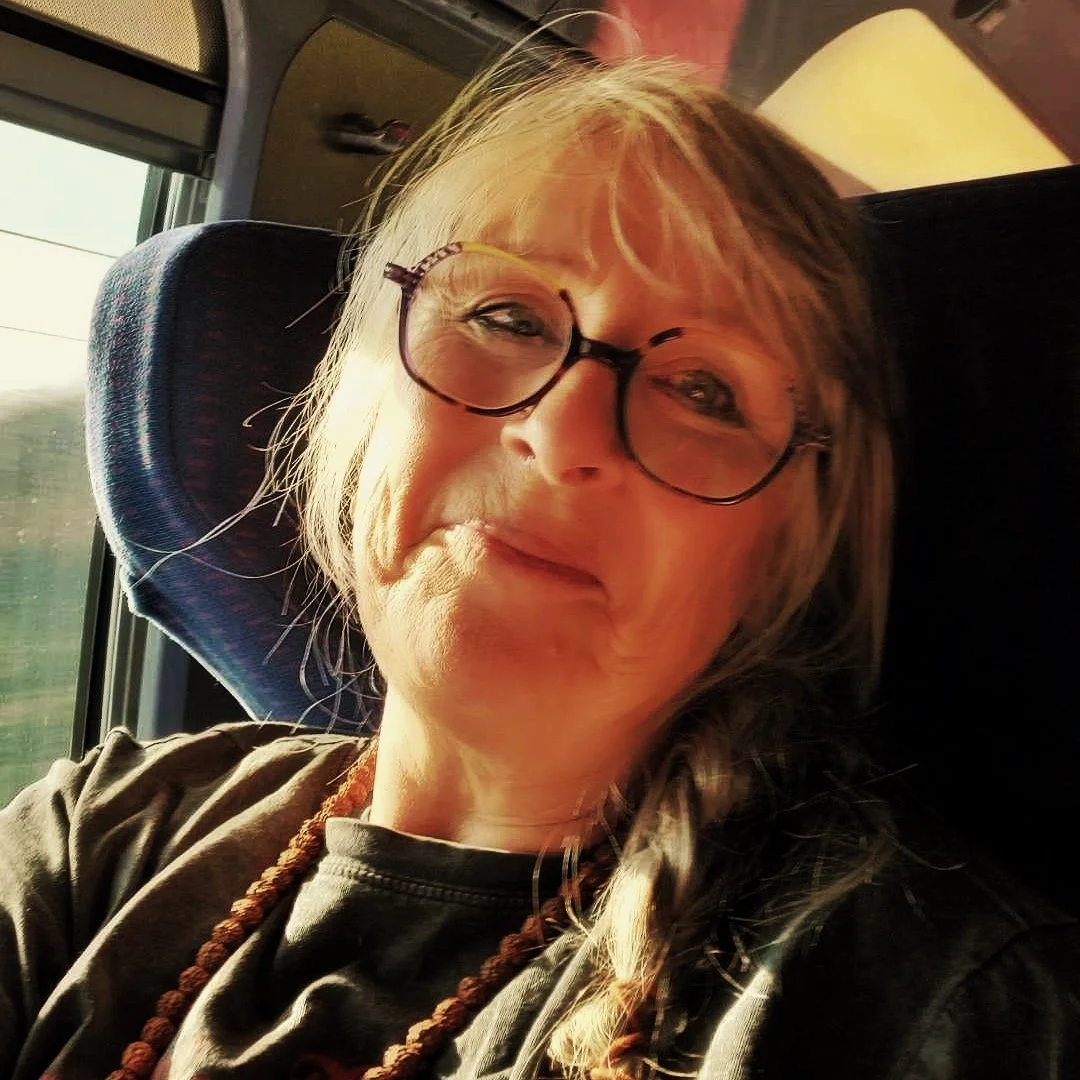 A woman with glasses and gray hair sitting on a bus, taking a selfie with natural light from the window illuminating her face.