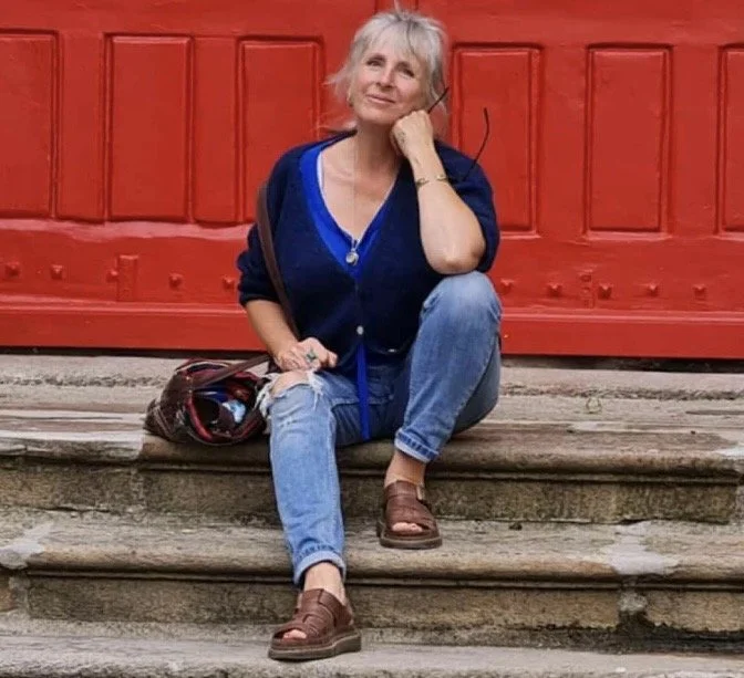 An older woman with short gray hair, wearing a dark blue jacket, blue shirt, and jeans, sitting on weathered steps in front of a red building, resting her chin on her hand and smiling.