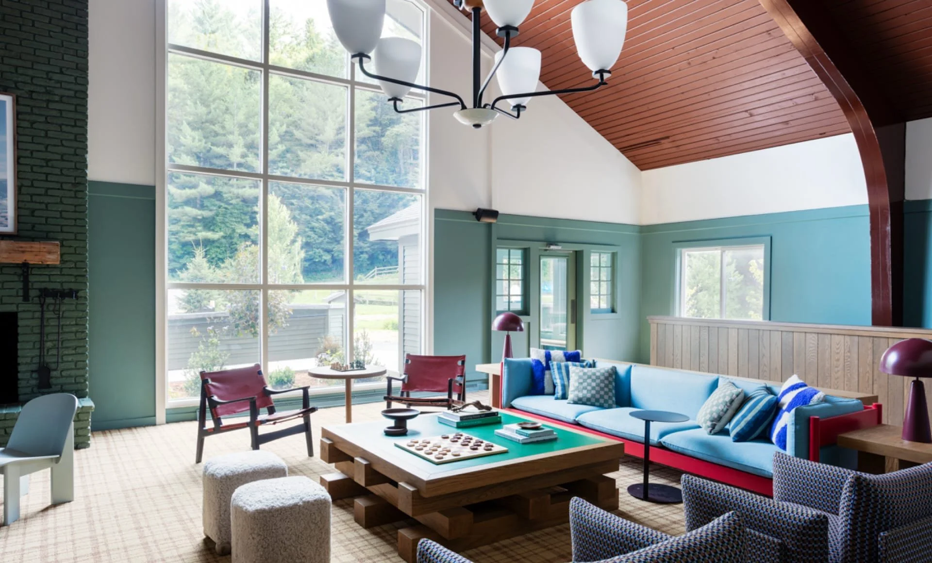 A bright living room with large corner windows showing a view of trees outside. The room has a high wooden ceiling with a modern chandelier. There is a turquoise sofa with various pillows, a wooden coffee table with board games, and several chairs and ottomans around. A green door and window are visible in the background.