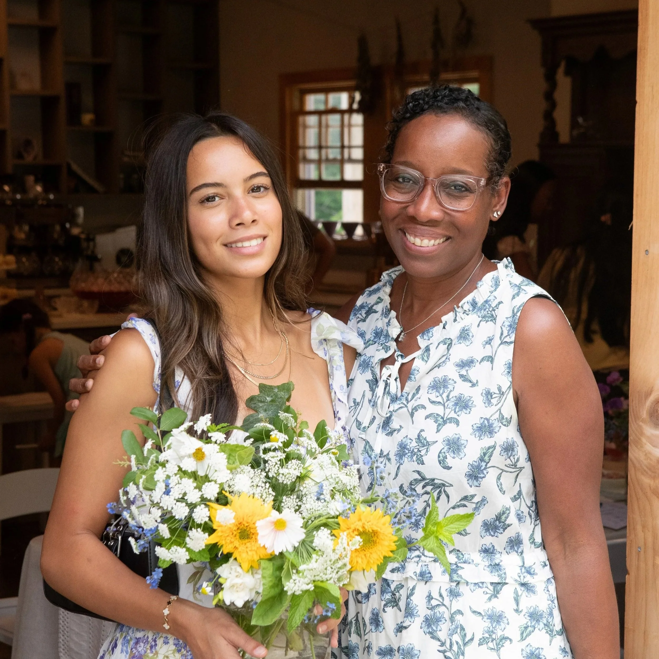 Mom and daughter smiling, one holding a bouquet of flowers, standing just outside farmhouse barn.
