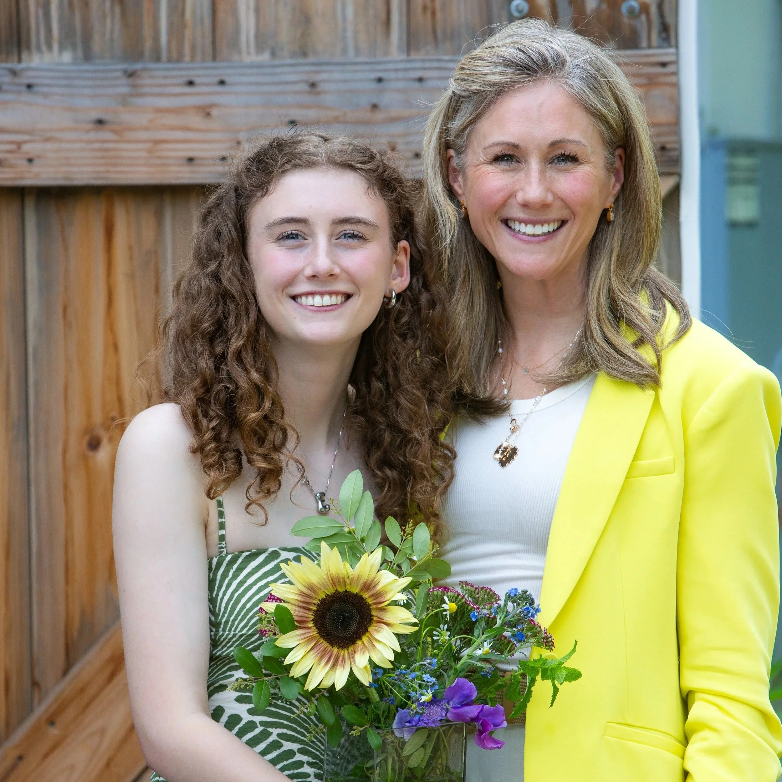 A woman and a girl smiling, holding a colorful bouquet of sunflowers, purple, and blue flowers, stands in front of a wooden fence.