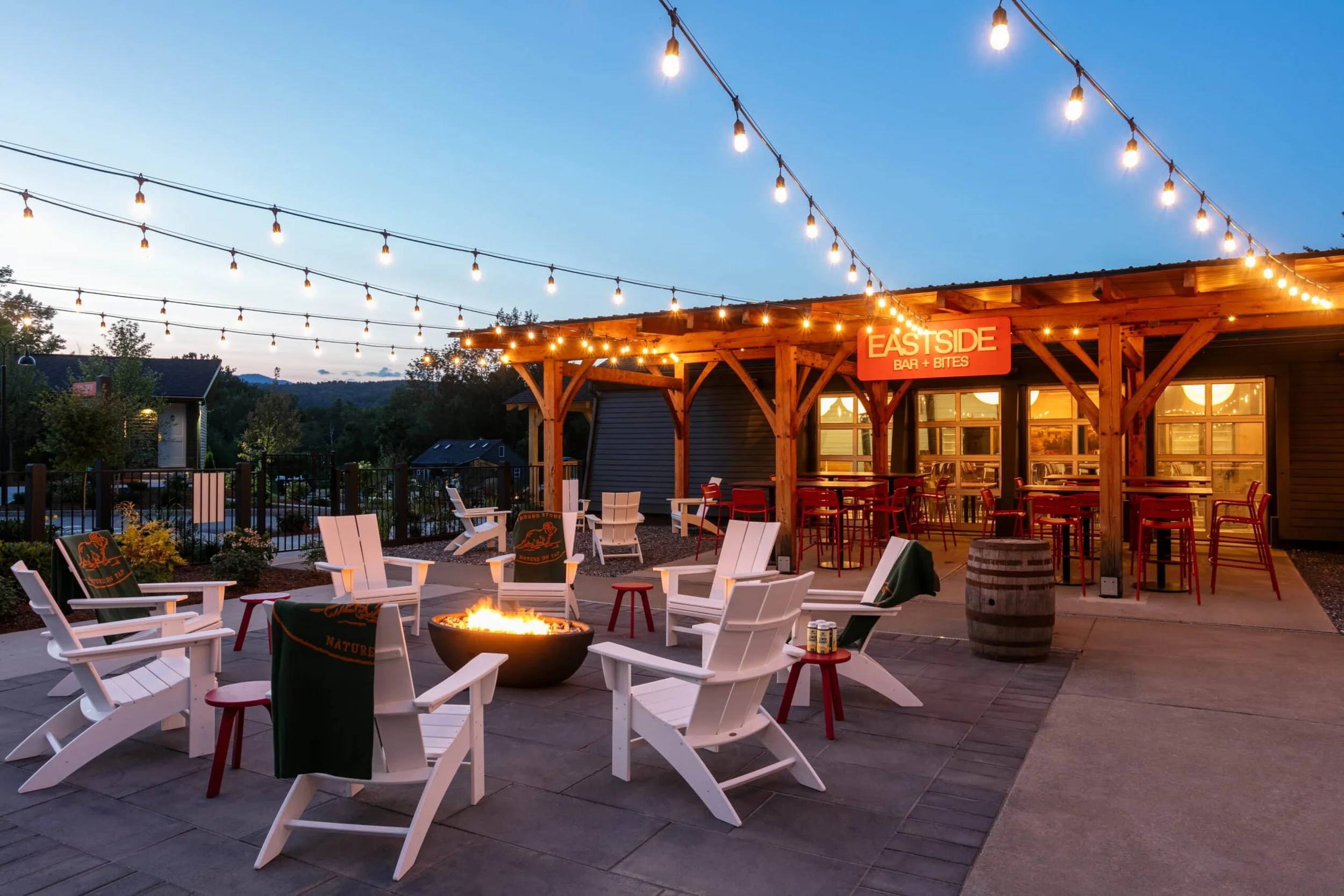An outdoor dining area at dusk with string lights overhead, white Adirondack chairs surrounding a fire pit, and a wooden building with a sign reading 'Eastside Bar + Bites' in the background.