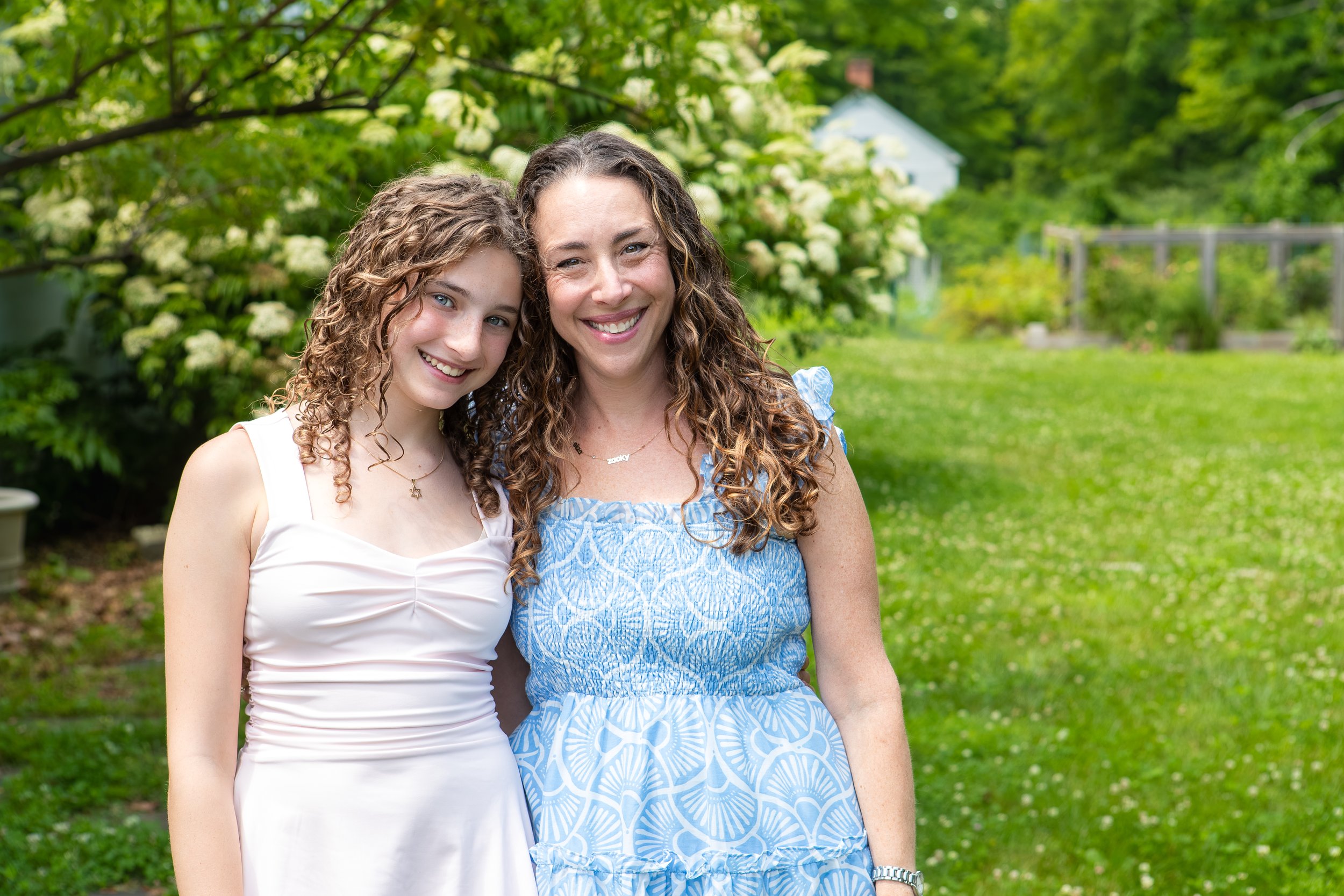 Mom and daughter standing outdoors in front of green trees and white flowers, smiling at the camera. A special moment that matters.