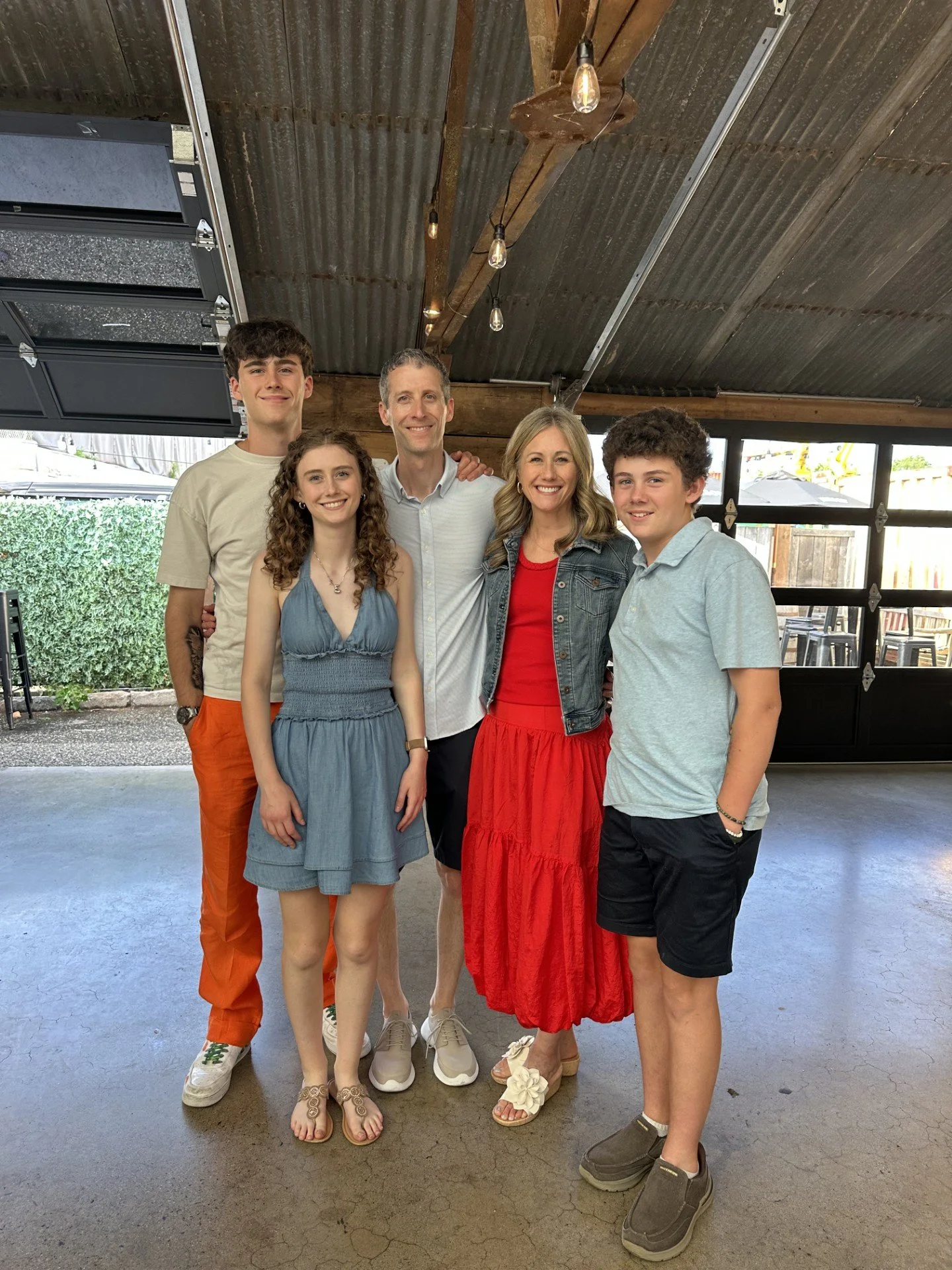 A group of six people standing together under a metal roof, smiling at the camera. The group includes three men and three women of varying ages, dressed casually. The background shows some outdoor elements like bushes and a wooden fence.