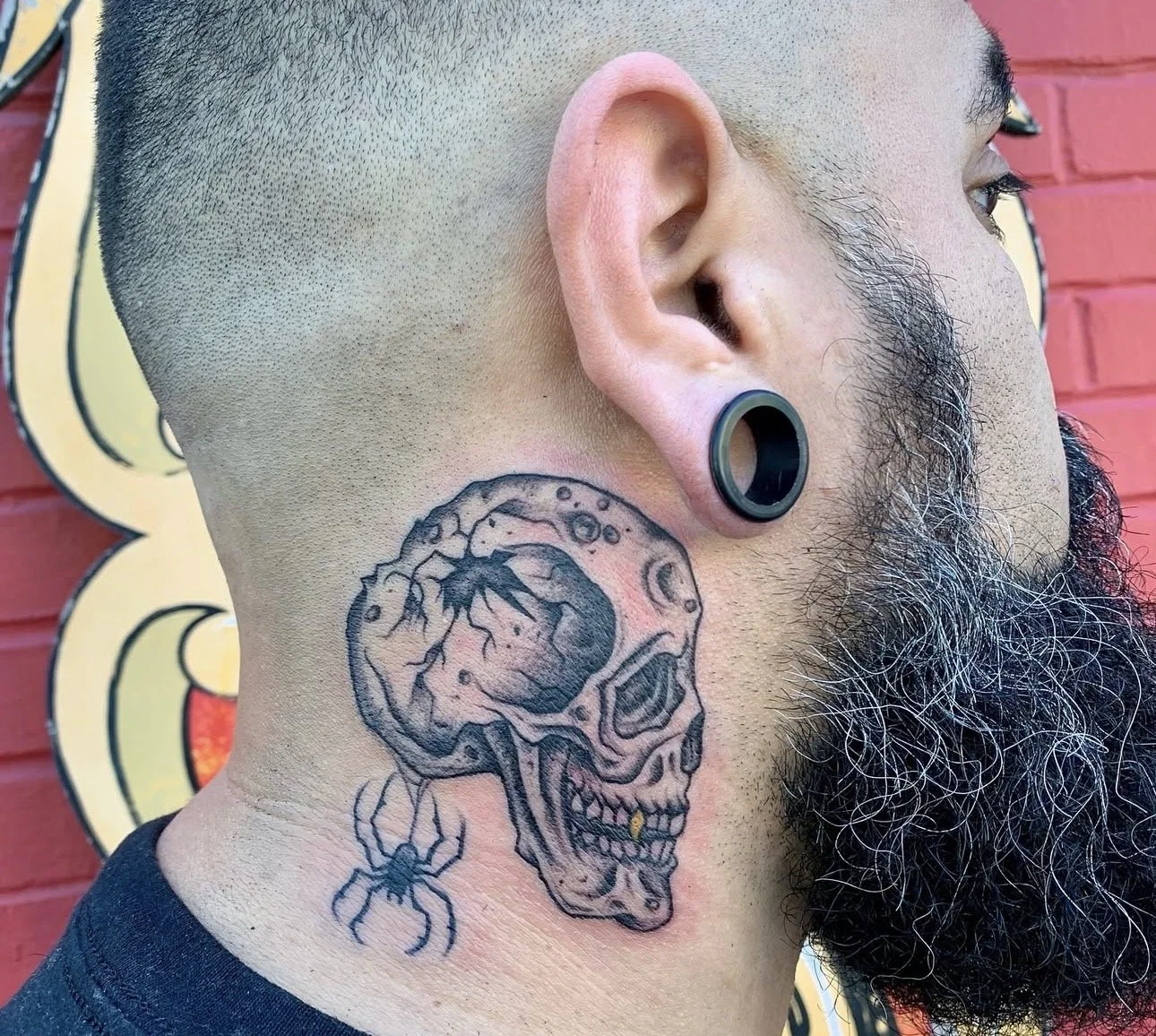 A man with a beard, large ear gauge, and a tattoo of a skull with a brain and spider on his neck.