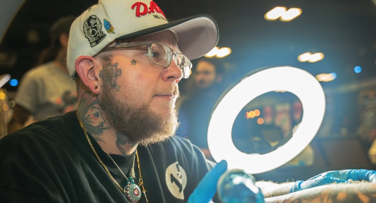 A man with tattoos, glasses, and a beard wearing a white cap with patches and a black T-shirt, sitting at a tattoo station, illuminated by a large circular light.