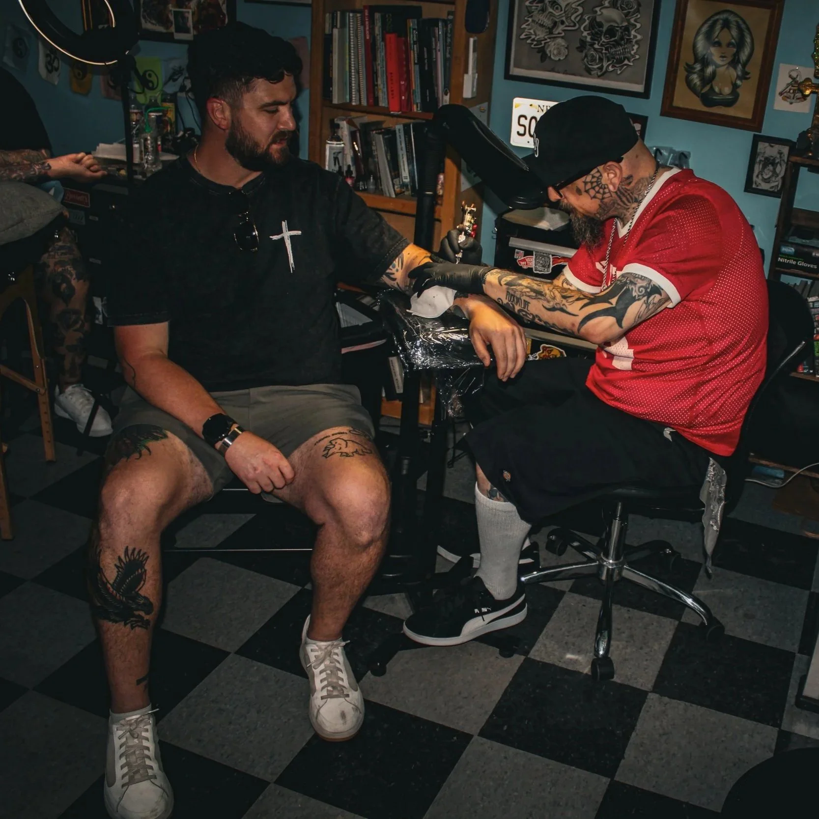 A man with tattoos on his legs and arms is sitting in a tattoo parlor, receiving a tattoo on his forearm from a tattoo artist. The man is wearing a black T-shirt, beige shorts, and white sneakers. The tattoo artist, wearing a black cap, glasses, and a red sports jersey, is working with tattoo equipment. The room has bookshelves, framed artwork on the walls, and a checkered black-and-gray floor.