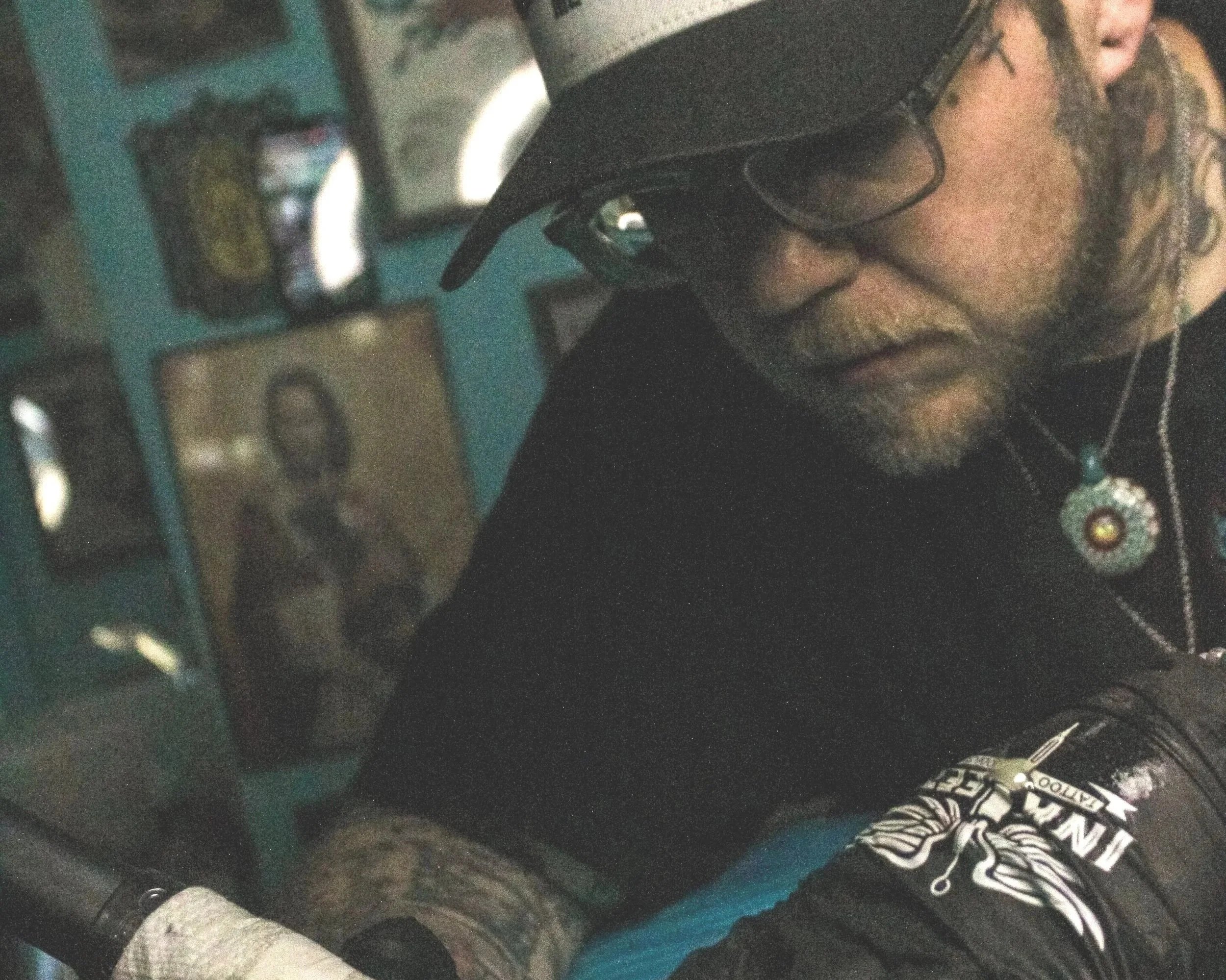 A person with glasses and curly hair wearing a black shirt, holding a teal-colored object, possibly a guitar, in a room with various objects in the background.