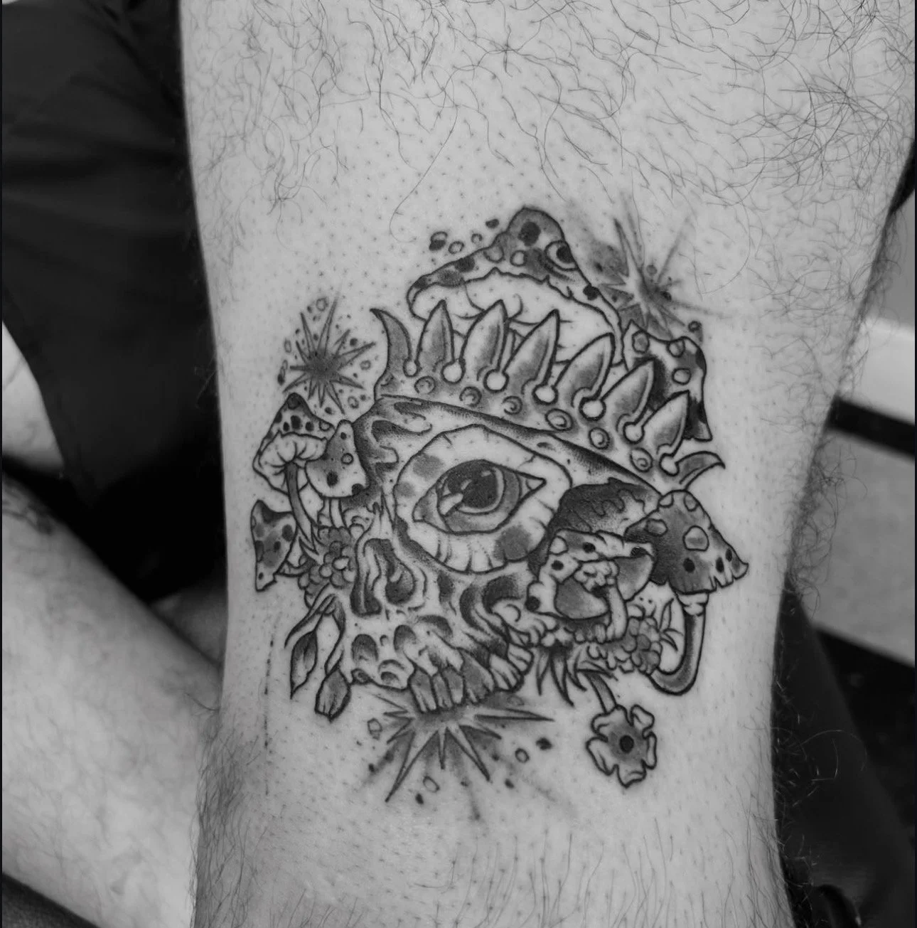 Tattoo of a detailed human skull with mushrooms, a large eye inside the skull, stars, and abstract designs.