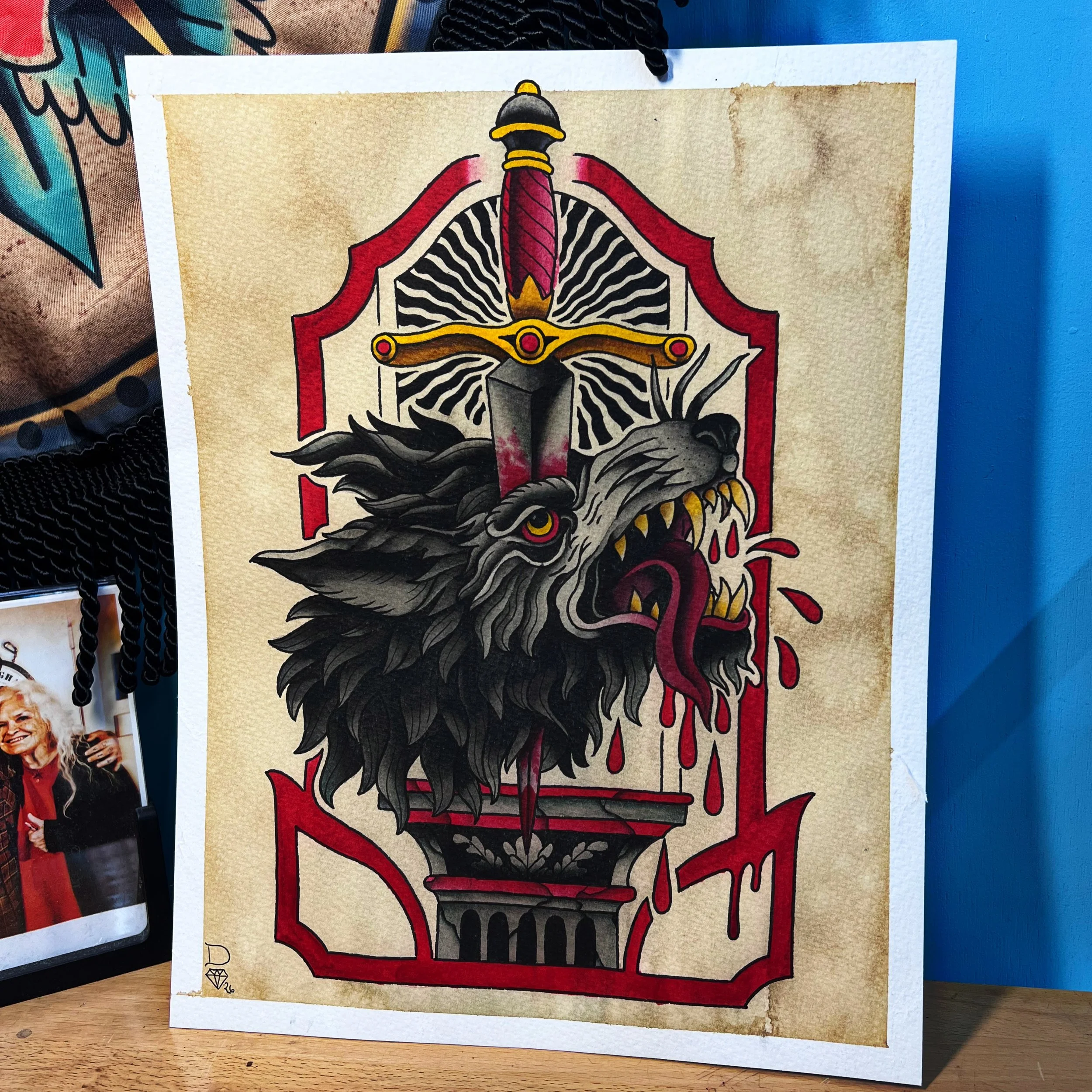 A colorful tattoo-style illustration featuring a fierce wolf with its mouth open, sharp teeth showing, and red eyes. The wolf's fur is detailed in shades of black and gray, and its expression is aggressive. The background has a red and black outline 