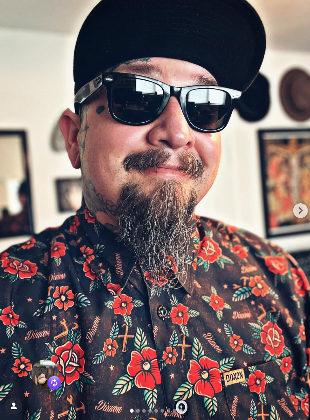 A man with a goatee and sunglasses taking a selfie. He is wearing a black hat and a floral shirt with red flowers and gold crosses.