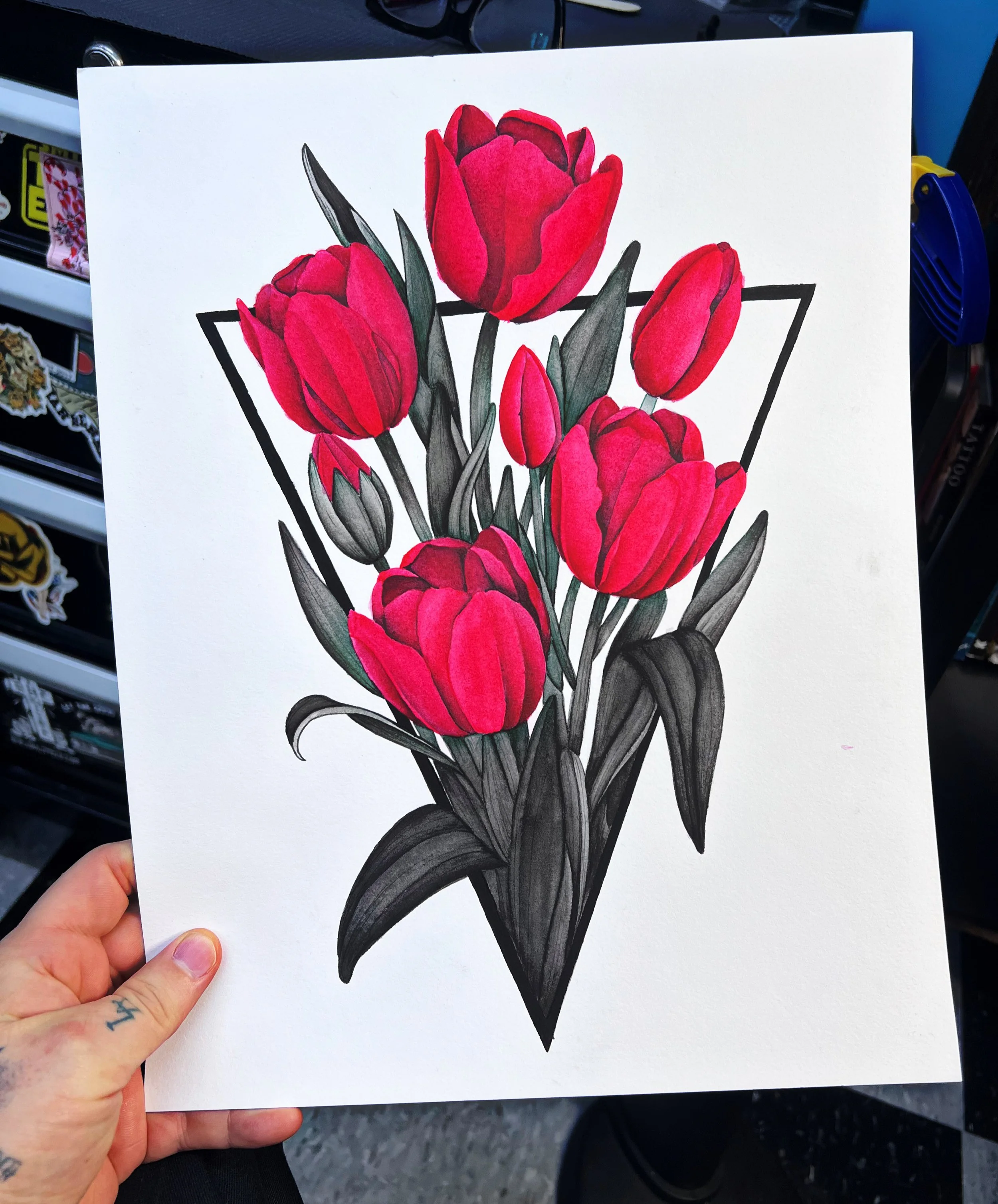 A person holding a white sheet of paper with a detailed colored pencil drawing of a bunch of red tulips with green leaves. The drawing is bordered with a black outline. In the background, there are shelves with various items, including stickers and other decorative objects.