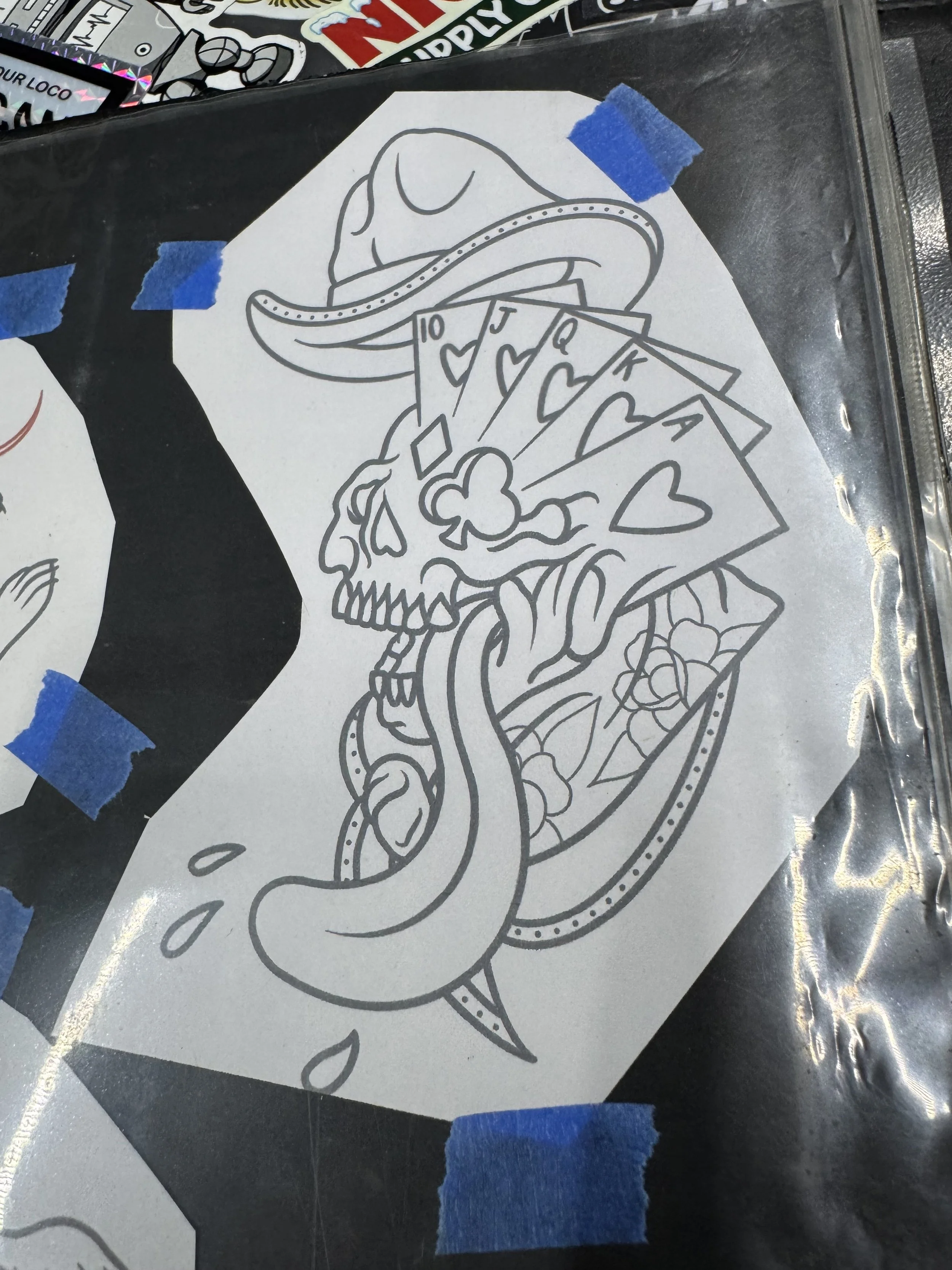 Black and white line drawing of a humorous hot dog with a skull, a baseball cap, and playing cards tucked into the bun, taped to a surface with blue painter's tape.