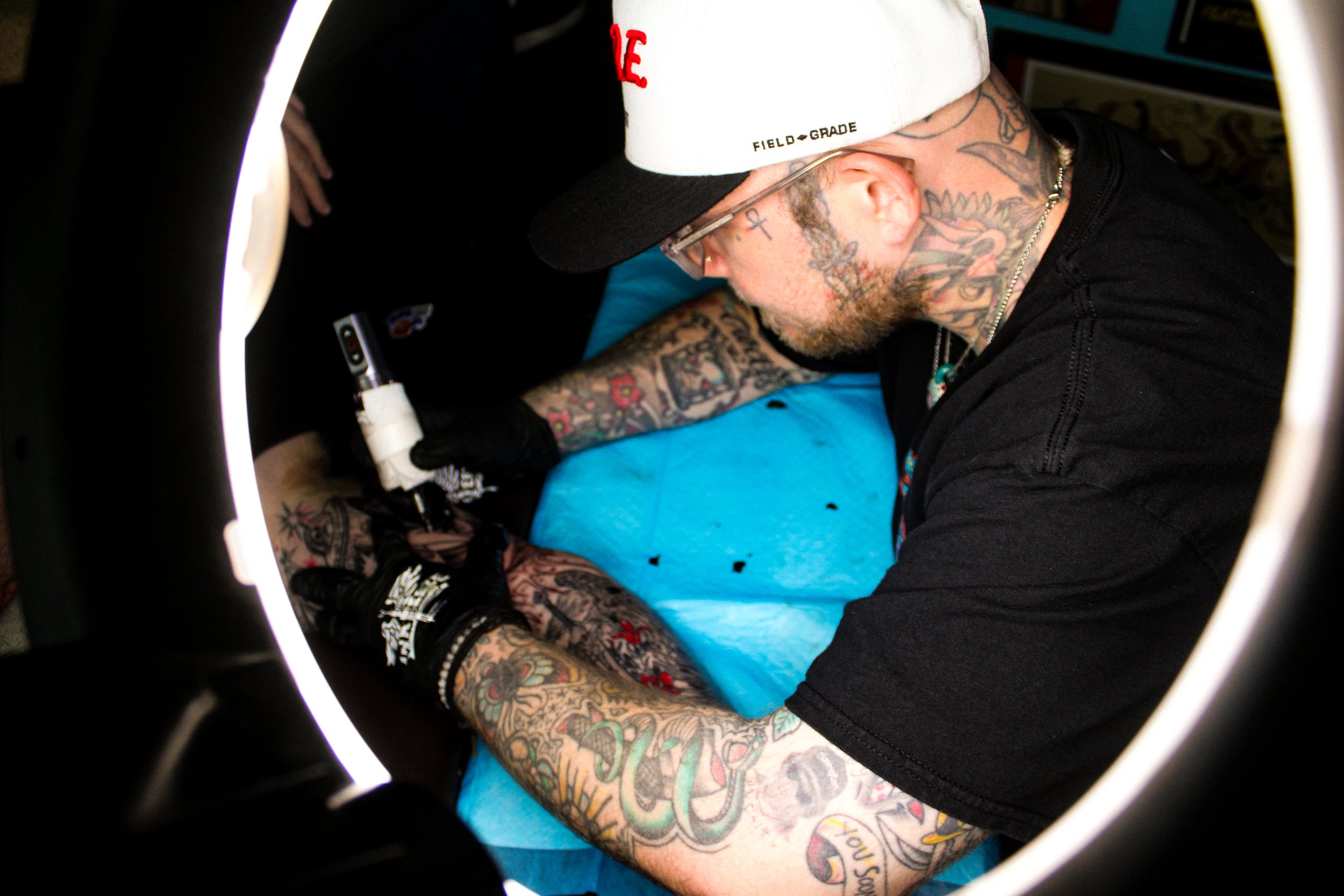 A person with extensive tattoos on their arms and neck, wearing glasses, a white and black baseball cap, and black clothing, appears to be getting a tattoo or performing a tattooing process. The image is taken through a ring light reflecting on a tattoo machine.