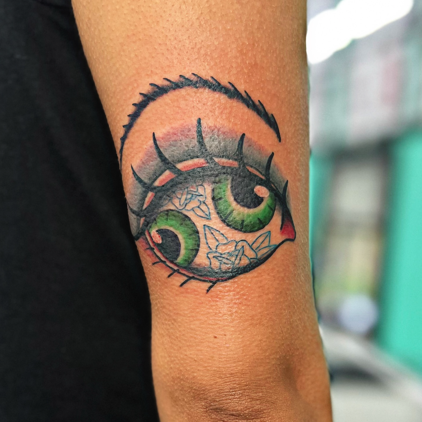 Tattoo of a stylized eye with green and black eyelashes on an individual’s arm.