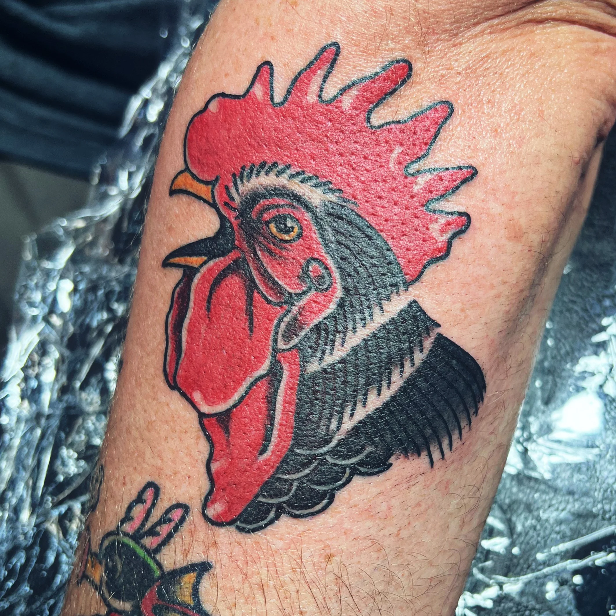 Close-up of a colorful tattoo on a person's skin, depicting a cartoonish red rooster with black outline and detailed features.
