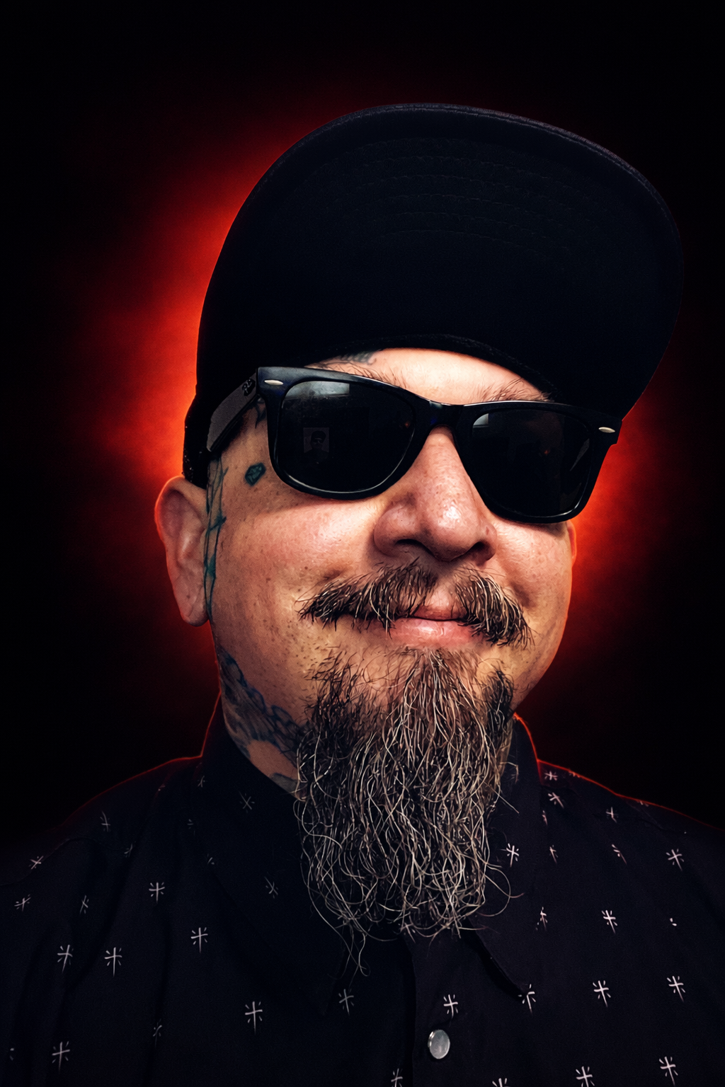 Close-up portrait of a man with sunglasses, a beard, and tattoos, wearing a black cap and black shirt, with a red halo glow behind him.