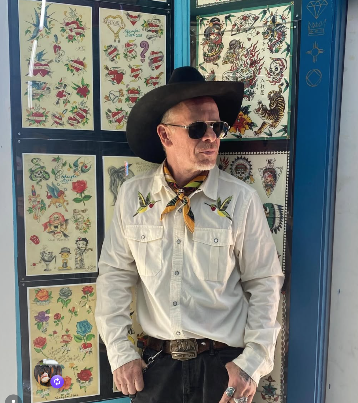 A man dressed in a Western-style outfit, wearing a black cowboy hat, sunglasses, a cream-colored shirt with decorative embroidery, a bandana around his neck, and standing in front of a display case filled with colorful traditional tattoo flash art.