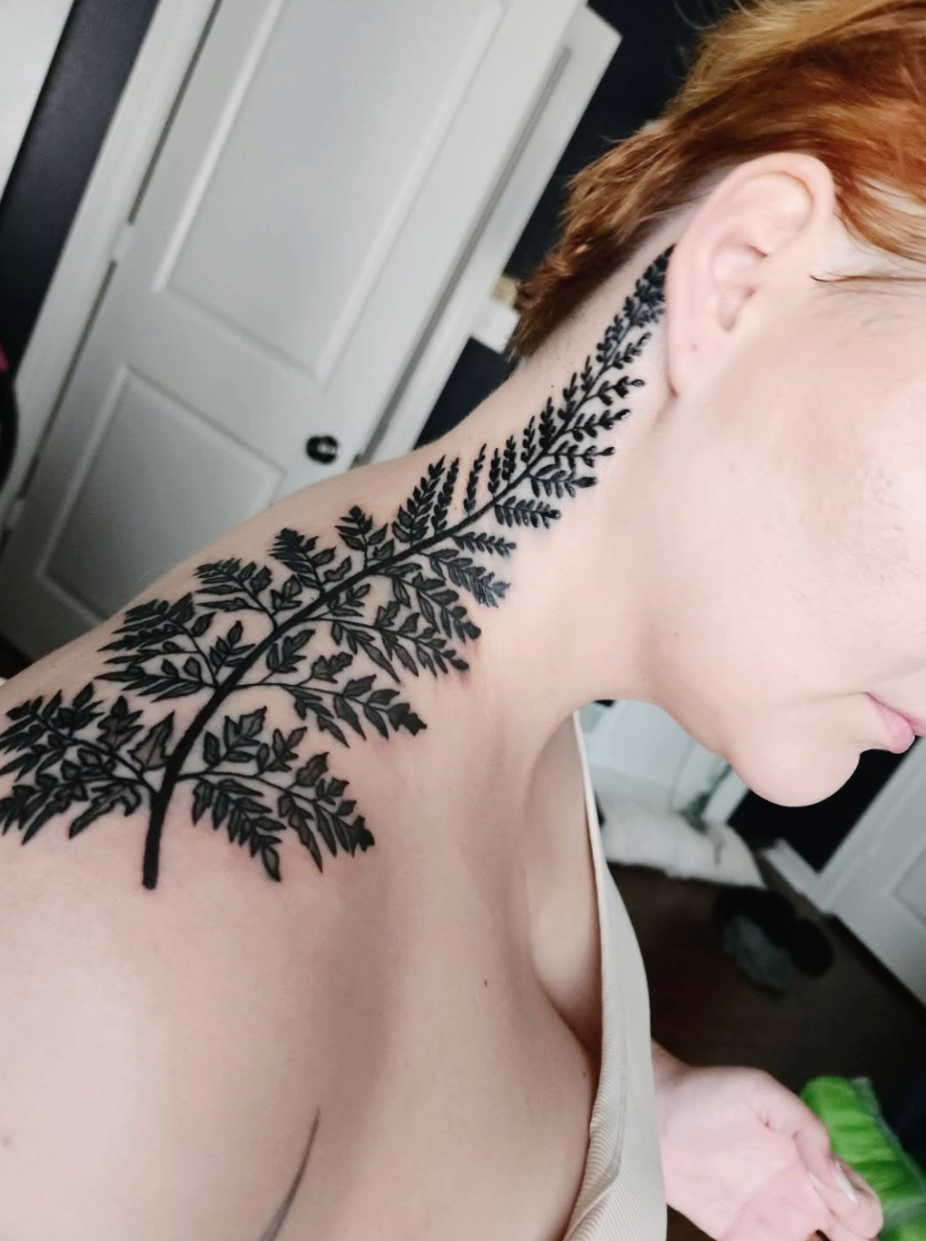 A woman with red hair is showing a large black fern tattoo on her shoulder and neck area.