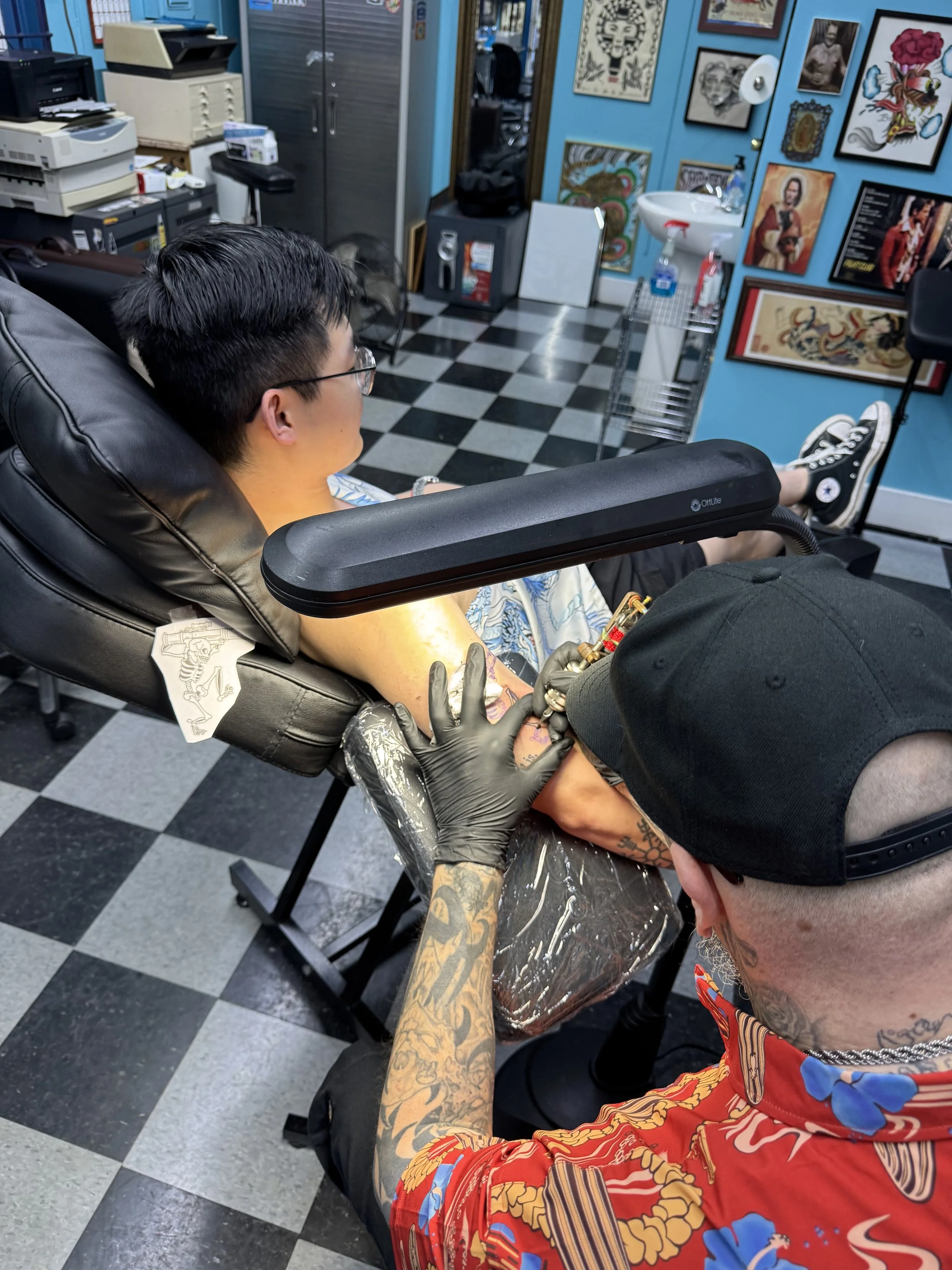 A man getting a tattoo on his upper arm in a tattoo studio. The man is wearing glasses and a floral shirt. The tattoo artist is wearing a black cap and gloves.