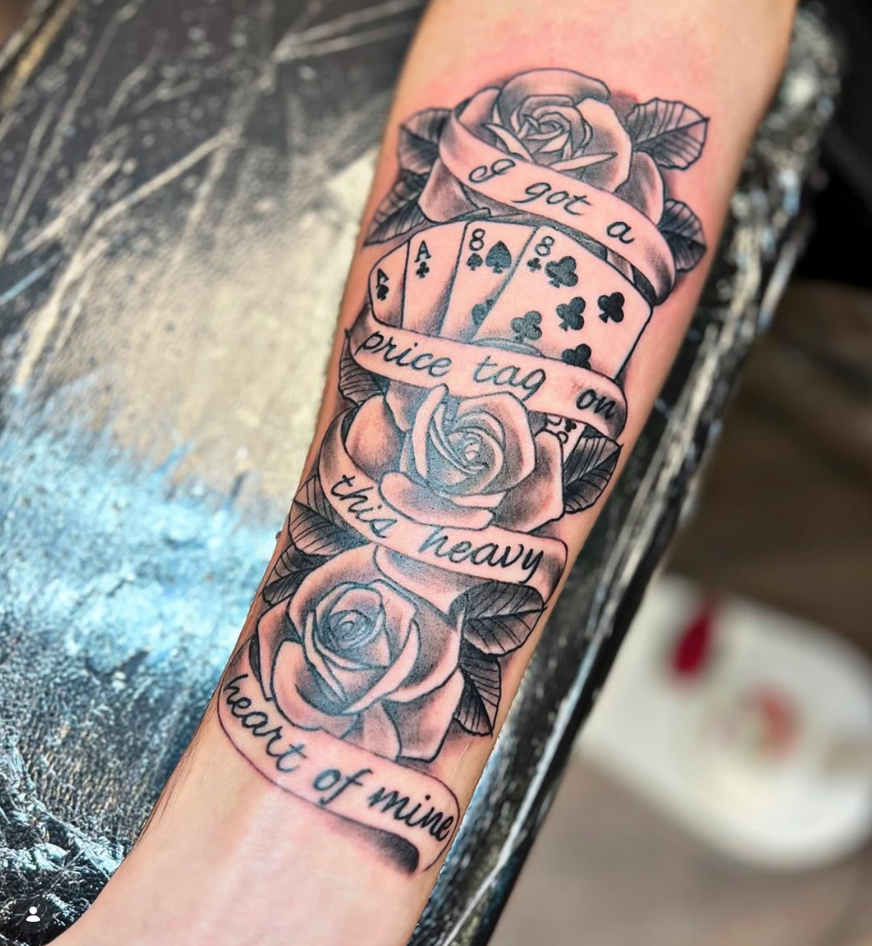 Black and gray tattoo on forearm featuring roses, playing cards, and ribbons with the text 'I'll 220 a price tag on this heavy heart'.